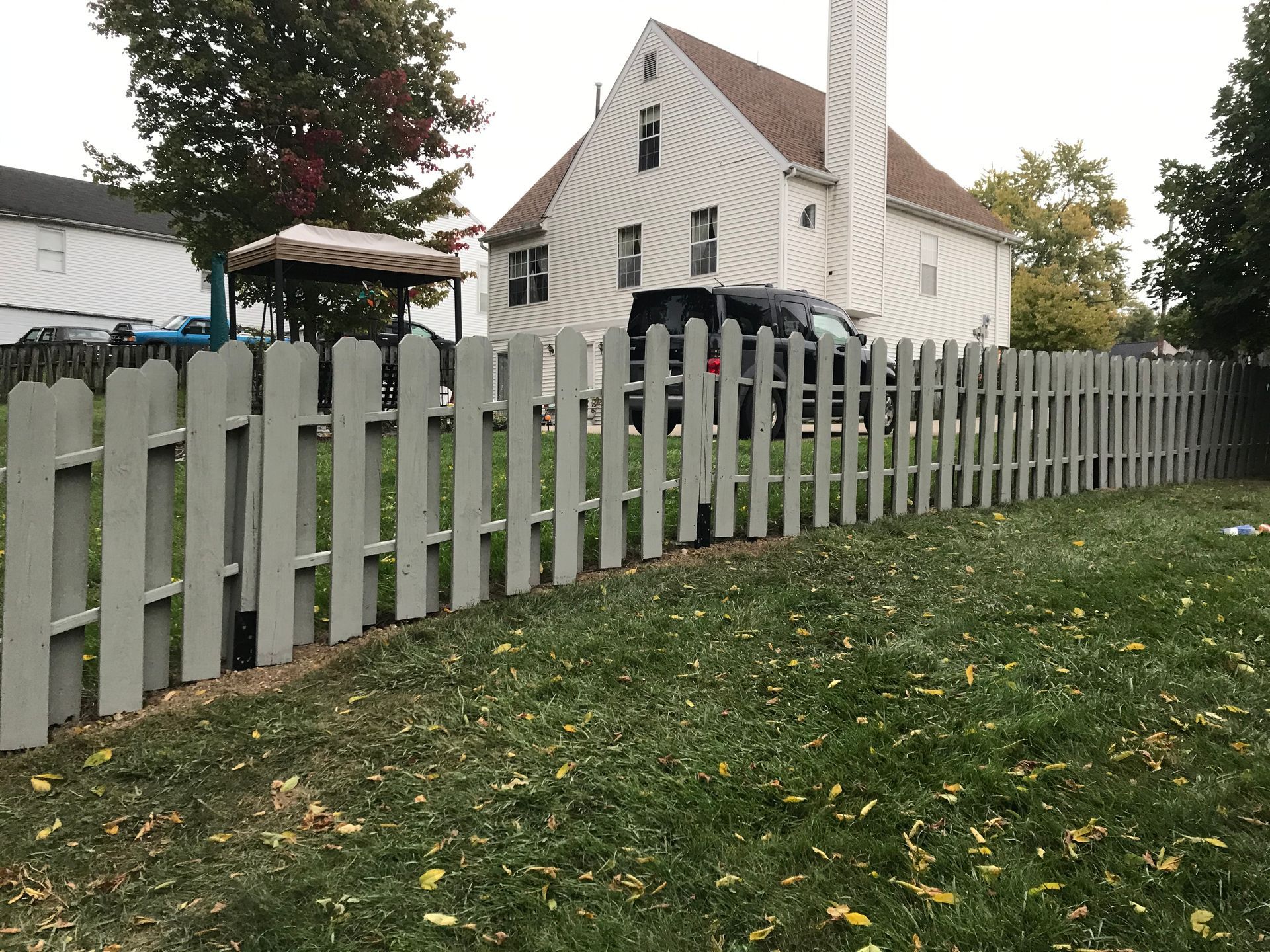 Gray picket fence in front of a two-story house with a parked black car. Green grass and leaves.