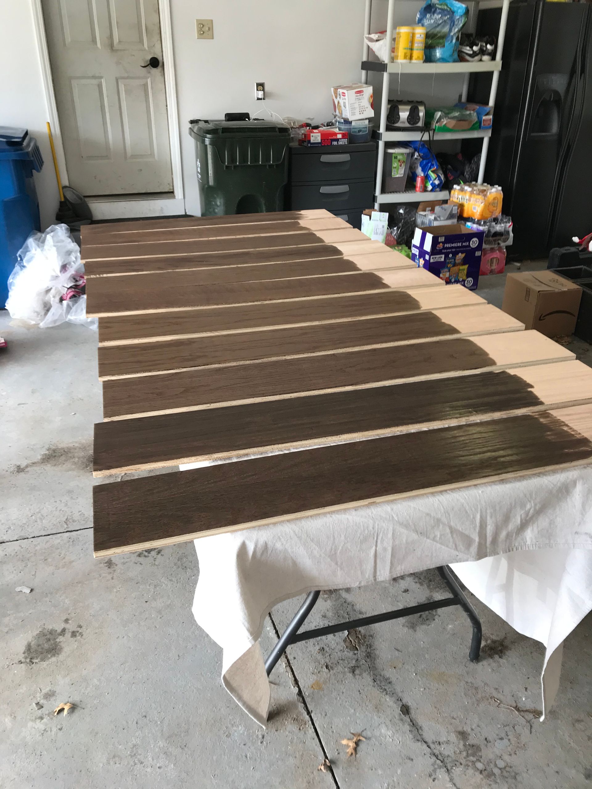 Wooden planks with varying shades of brown stain, laid on a white-covered table in a garage.
