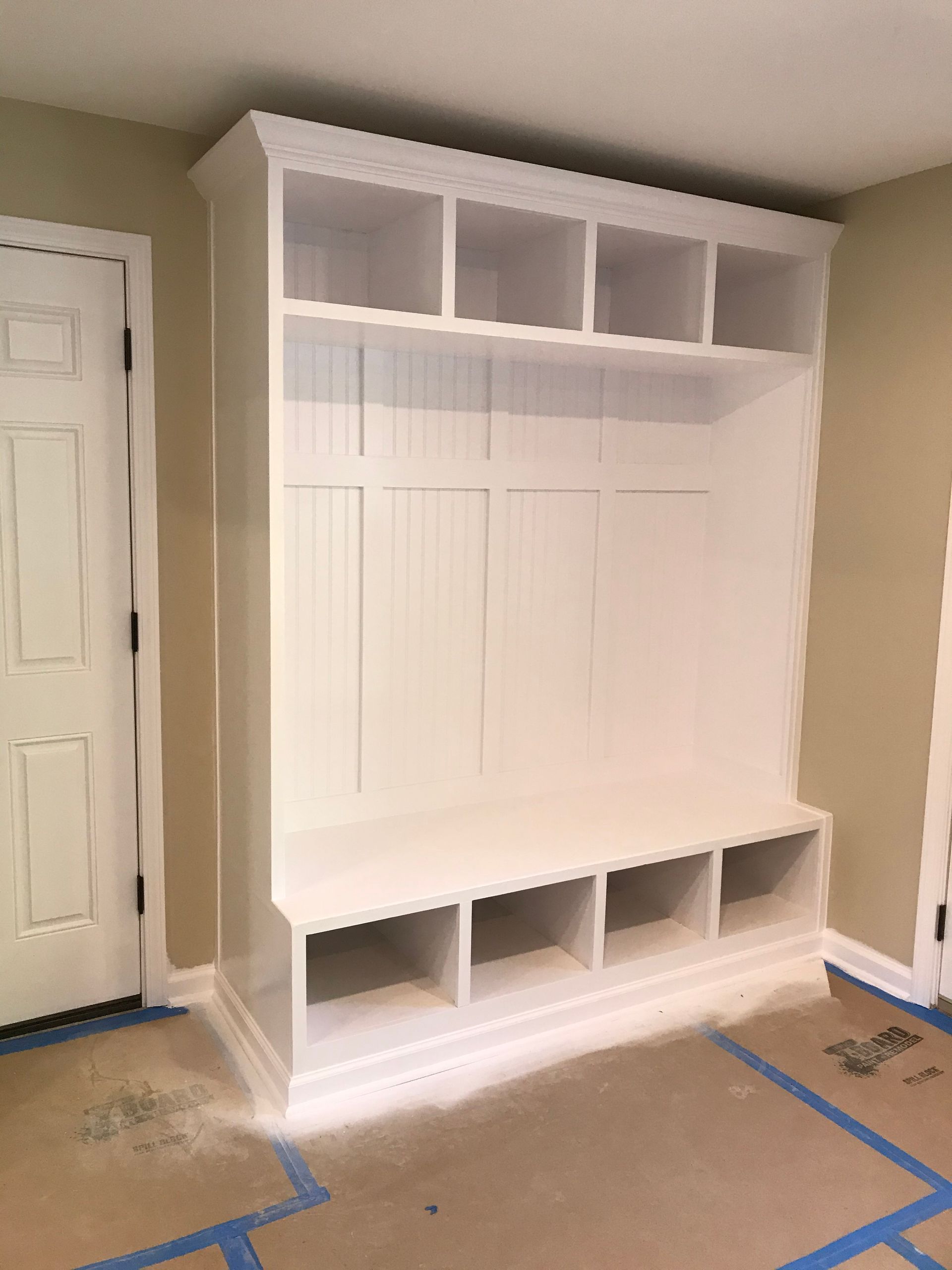 White built-in mudroom storage unit with bench, cubbies, and upper shelves, against beige walls.