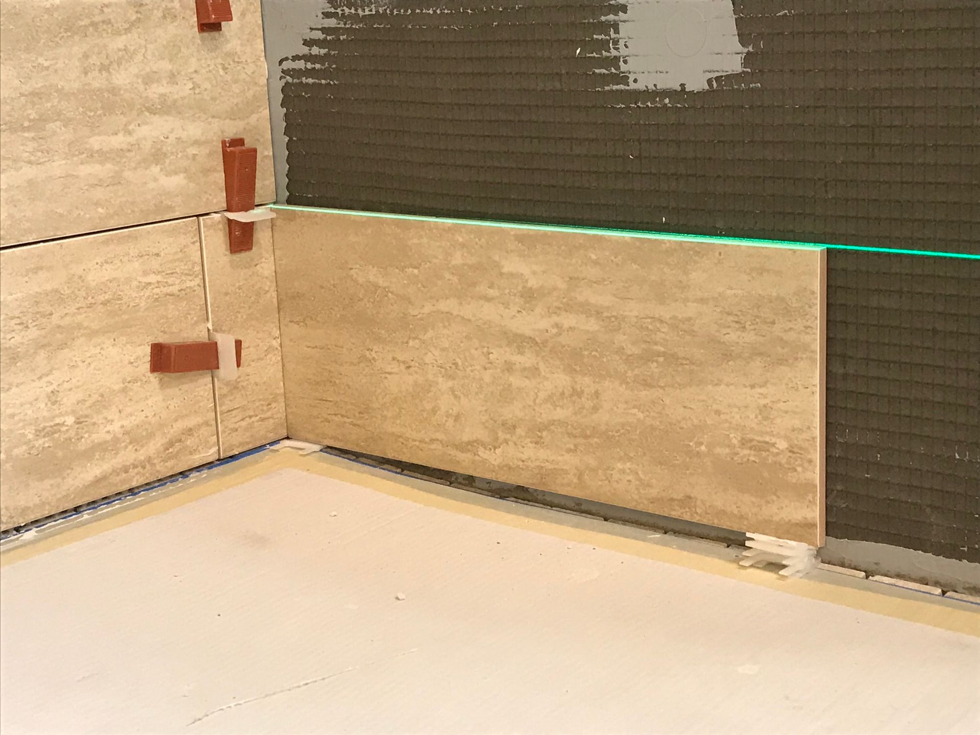 Tile installation in progress: Beige tiles on walls, with level lines and spacers.