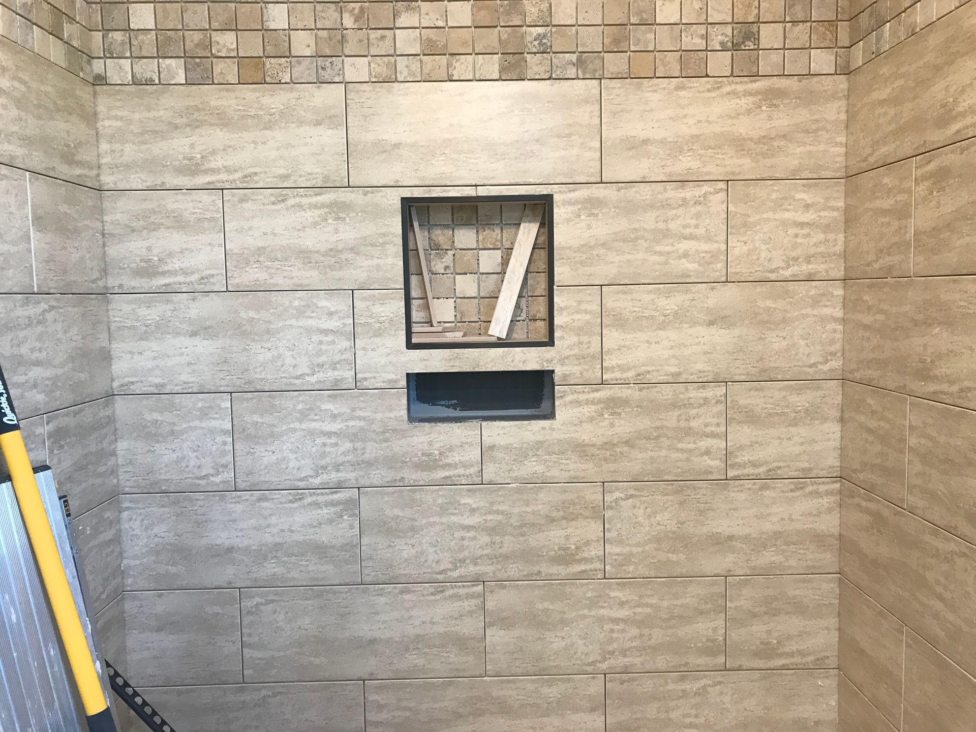 Shower wall with tan rectangular tiles, a tiled niche, and a mosaic border.