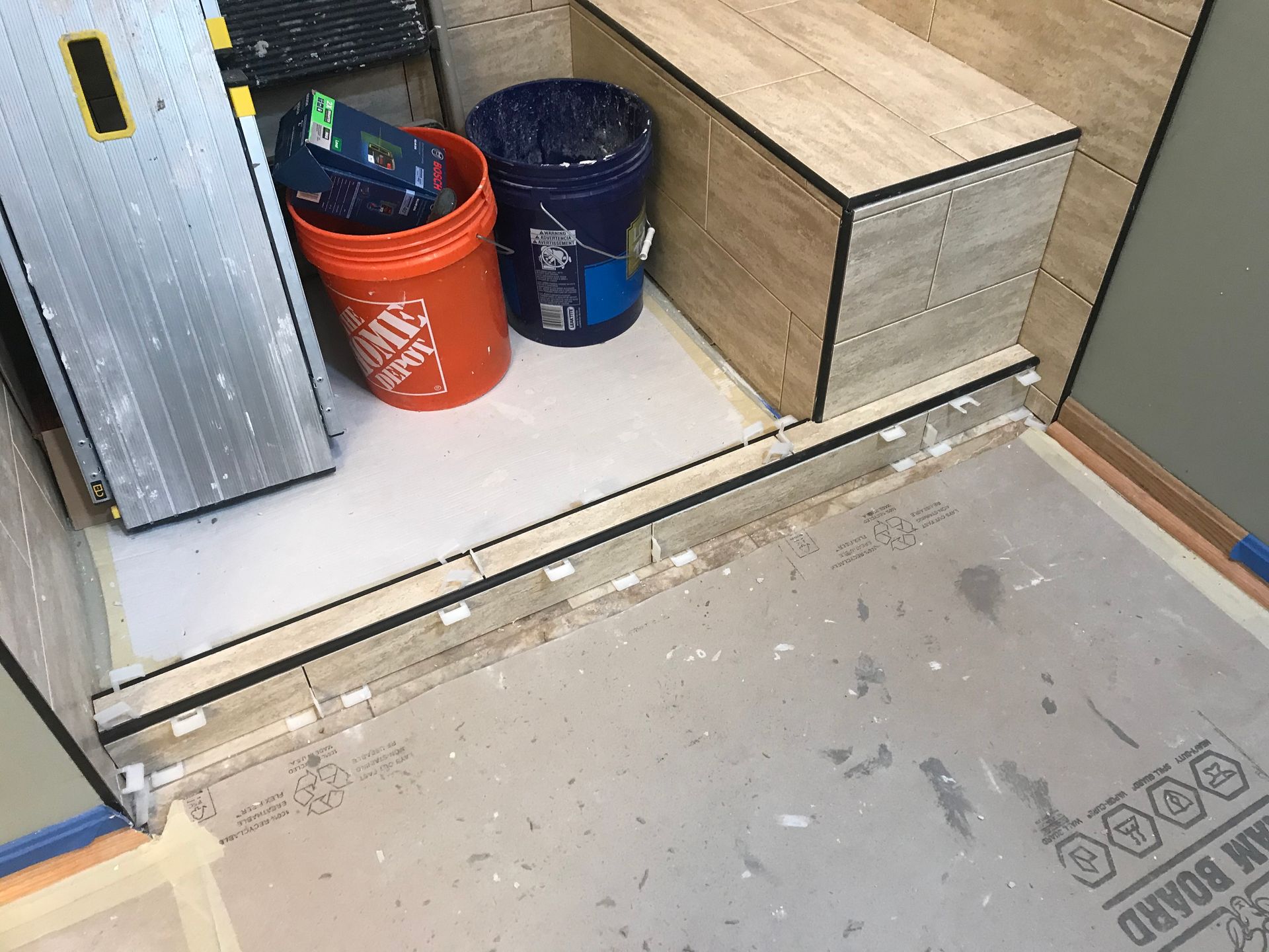 Shower tile installation in progress; tiles on floor and wall, tools, buckets, and bench present.