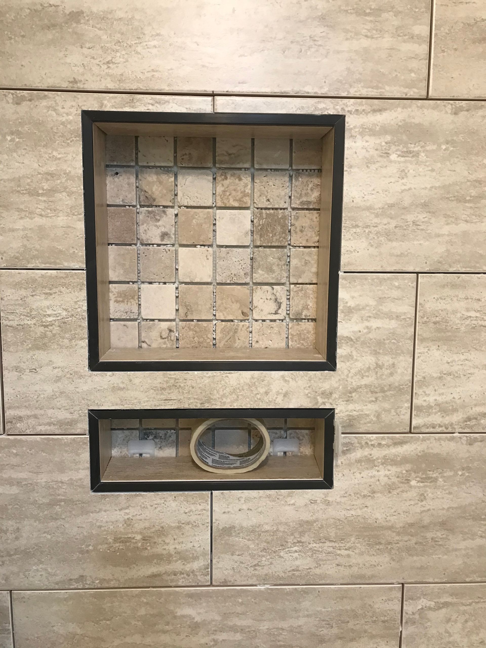 Recessed tiled shelf with a smaller shelf beneath, both with dark trim, in a tiled wall.