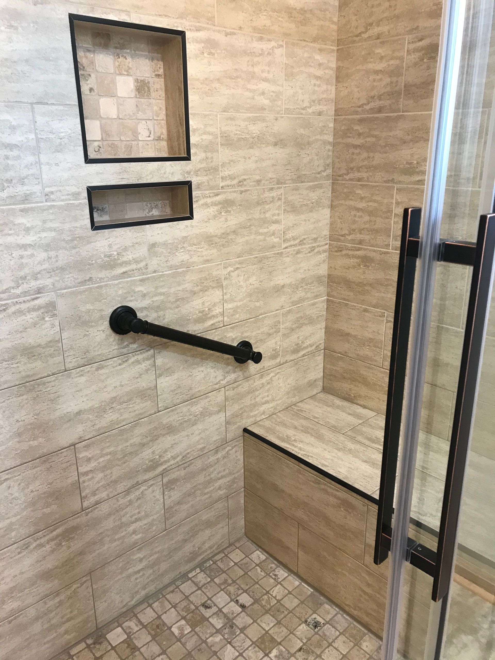Shower with beige tile, built-in bench, grab bar, and recessed shelving. Glass door on the right.