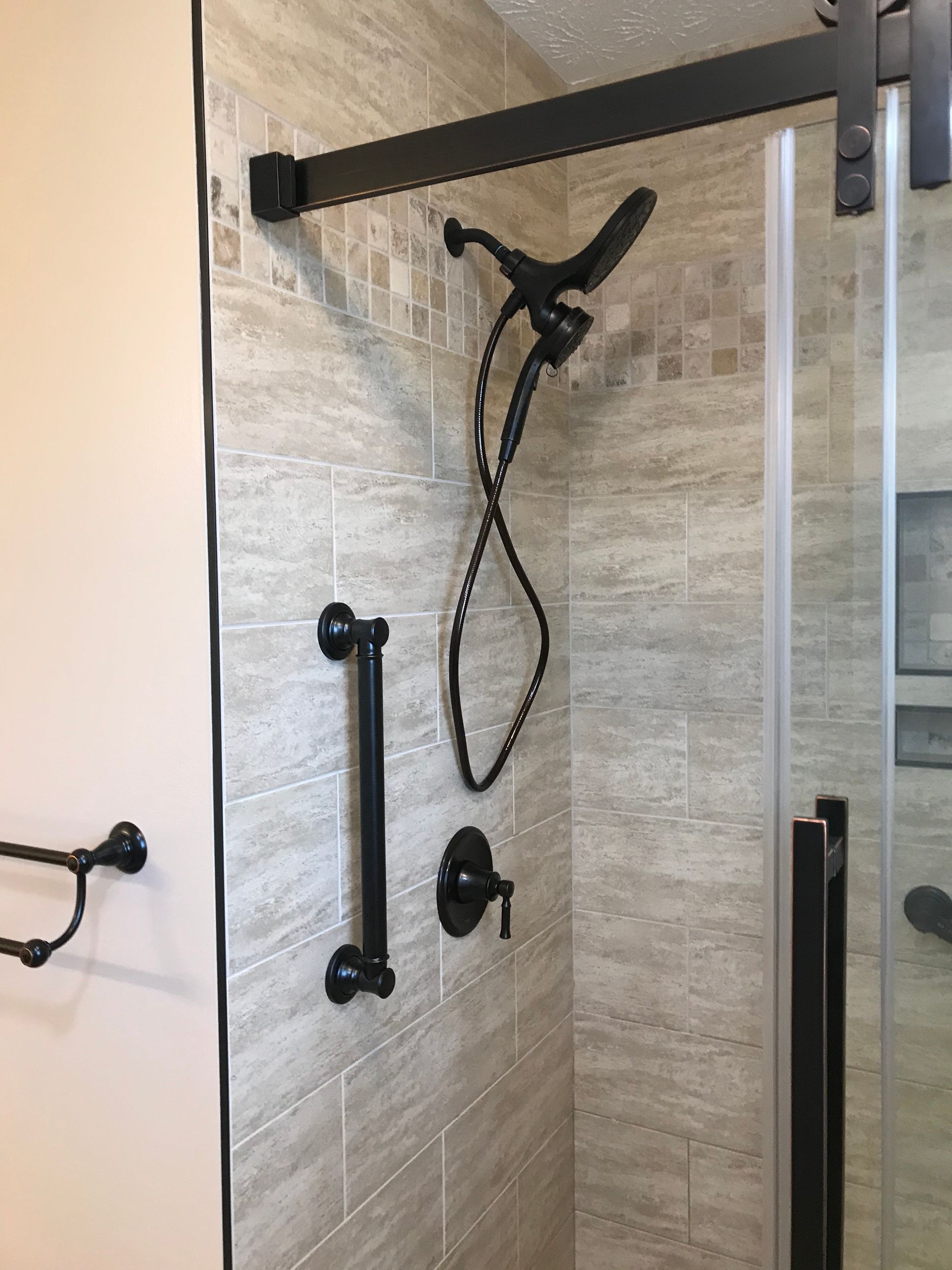 Shower with black fixtures and a grab bar on tile wall, next to a glass door.