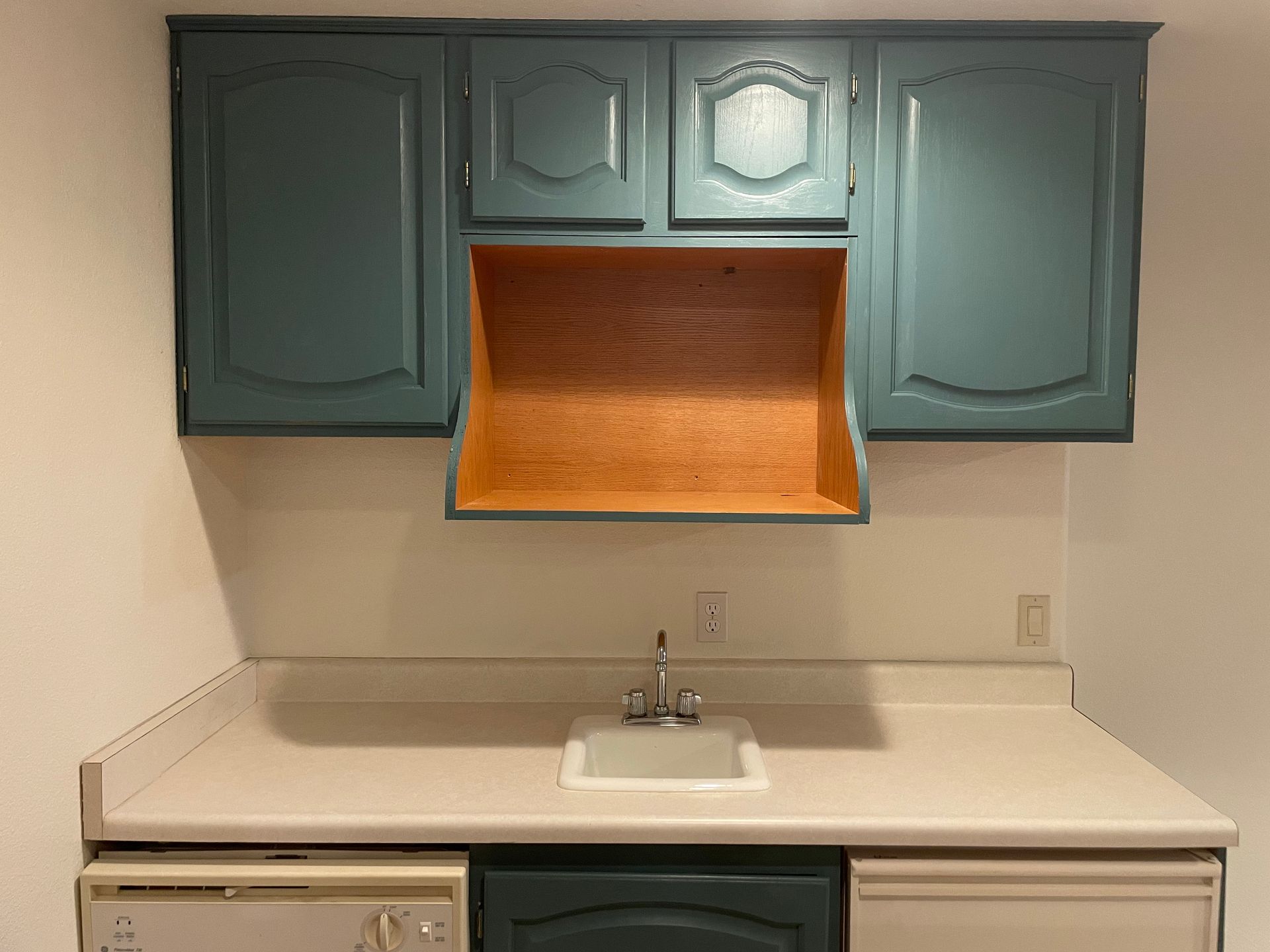 Kitchen cabinets and sink. Teal cabinets above a white countertop. Sink centered with a dishwasher on either side.
