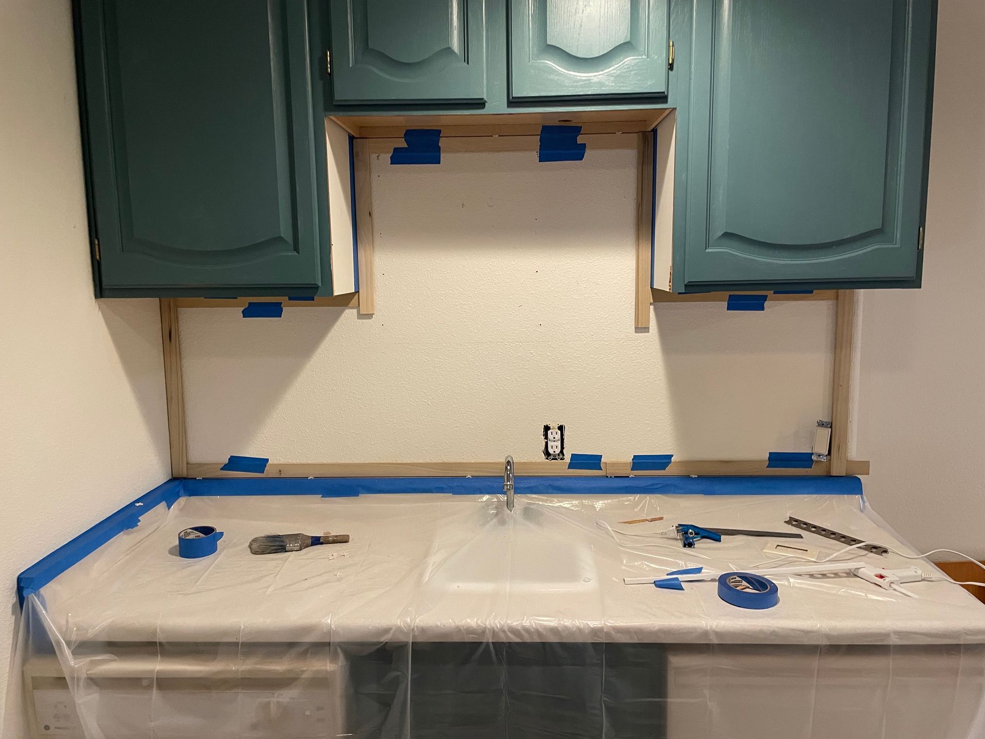 Kitchen backsplash installation in progress; cabinets painted teal, countertops covered, taped off.