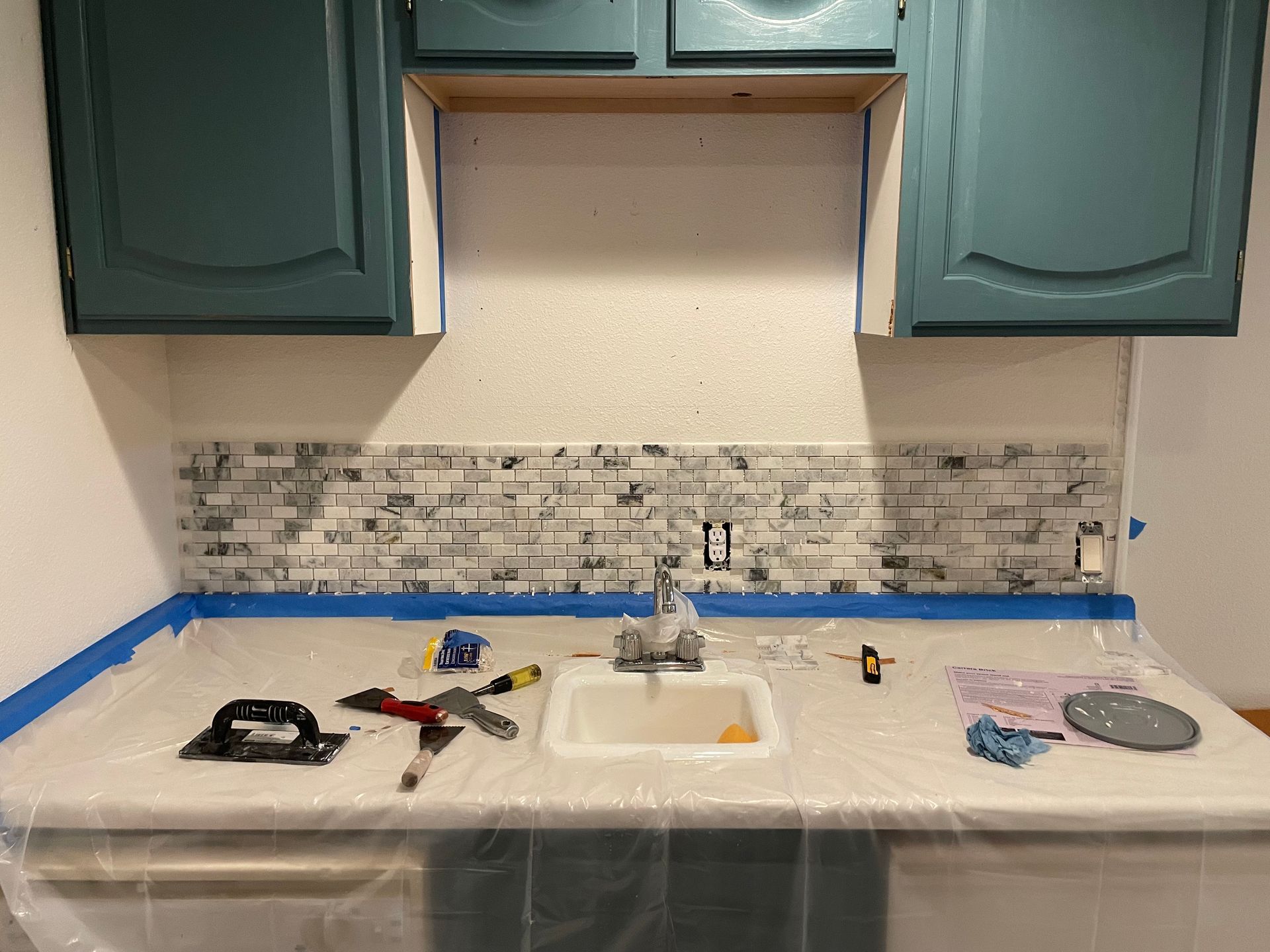 Kitchen backsplash tile installation in progress: grey and white mosaic tiles, blue cabinets.
