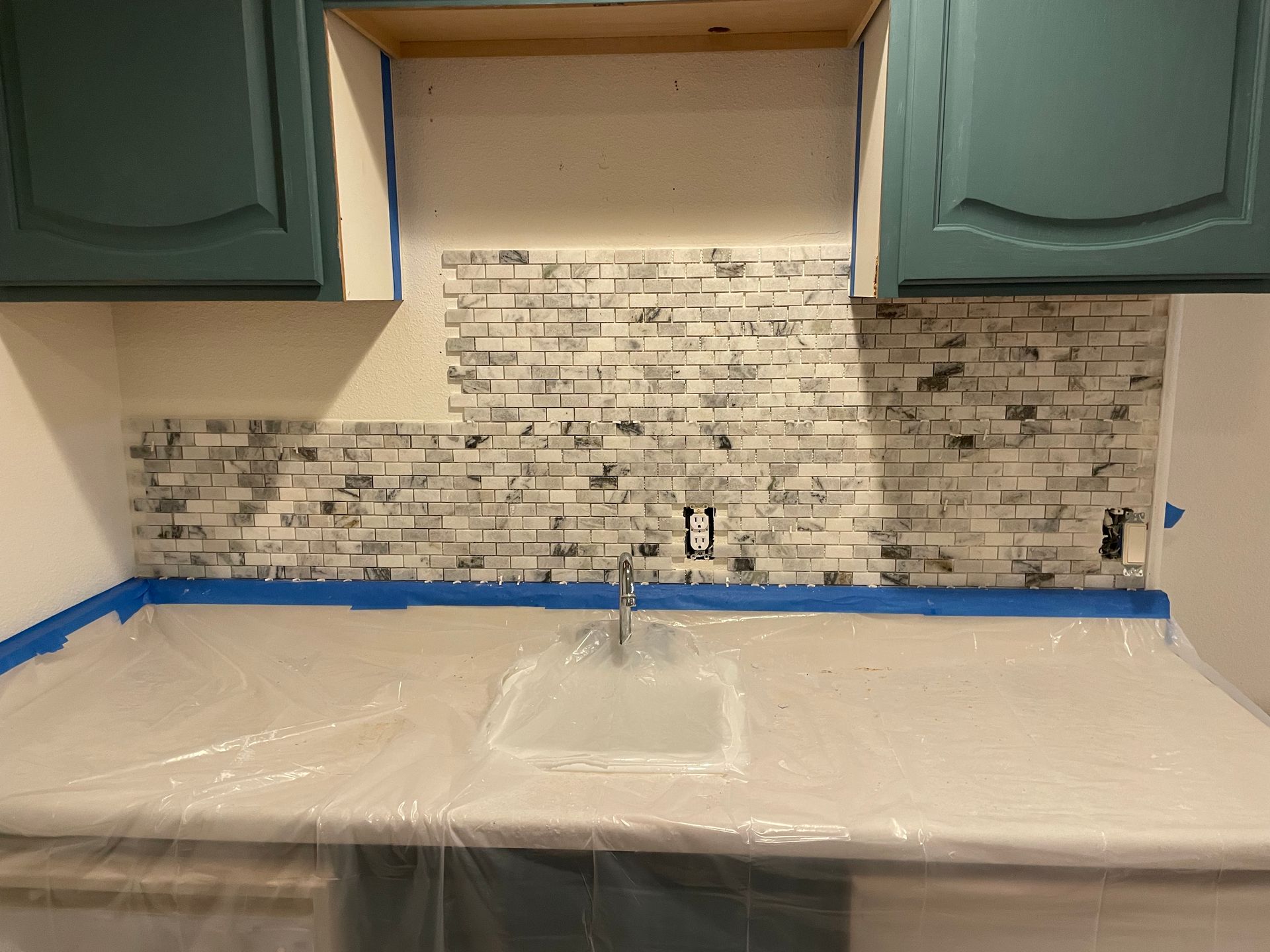 Kitchen backsplash under construction; white, gray, and black tile, blue taped counter, green cabinets.