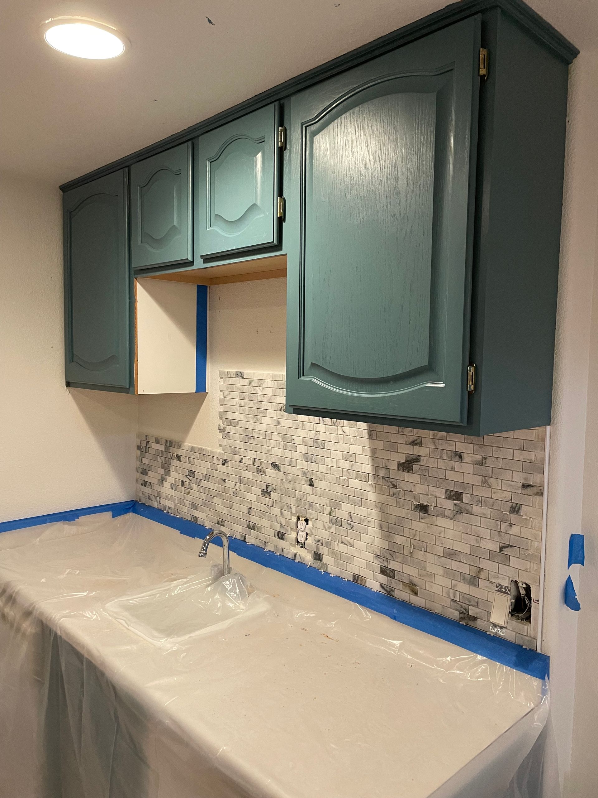 Teal kitchen cabinets above a white countertop and mosaic backsplash during renovation, taped off.