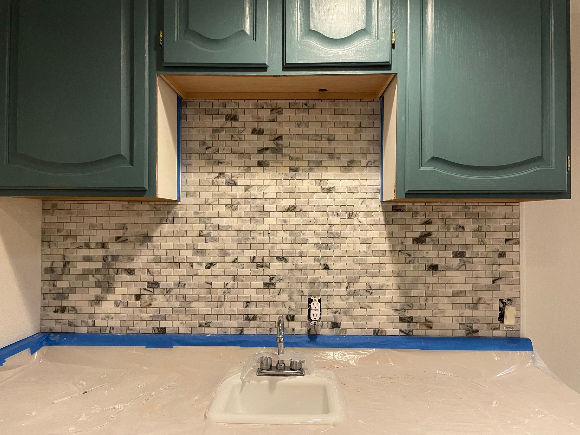 Kitchen backsplash installation in progress: teal cabinets, white and gray mosaic tiles, sink.