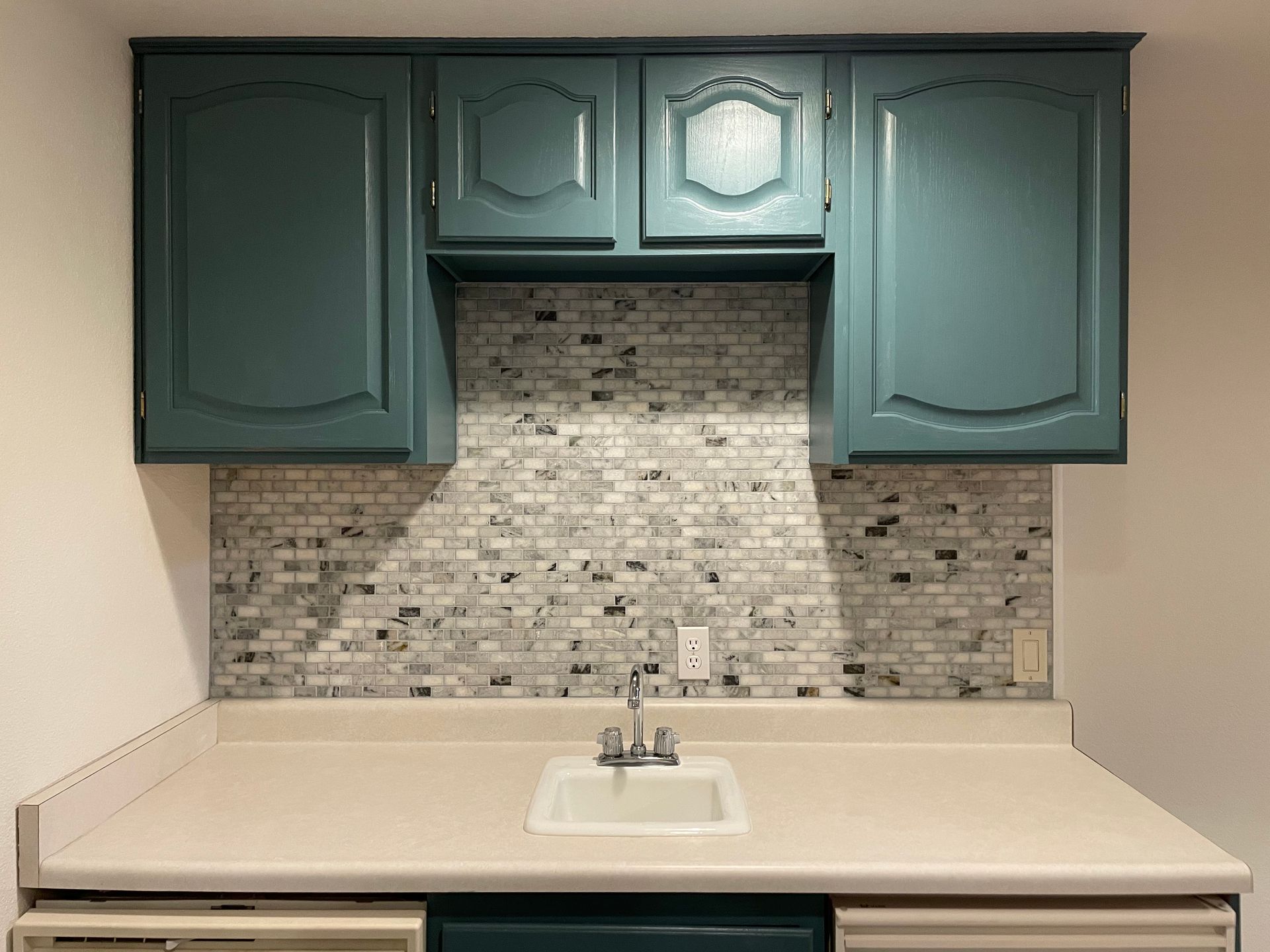 Kitchenette with teal cabinets, mosaic backsplash, and white countertop over a sink.