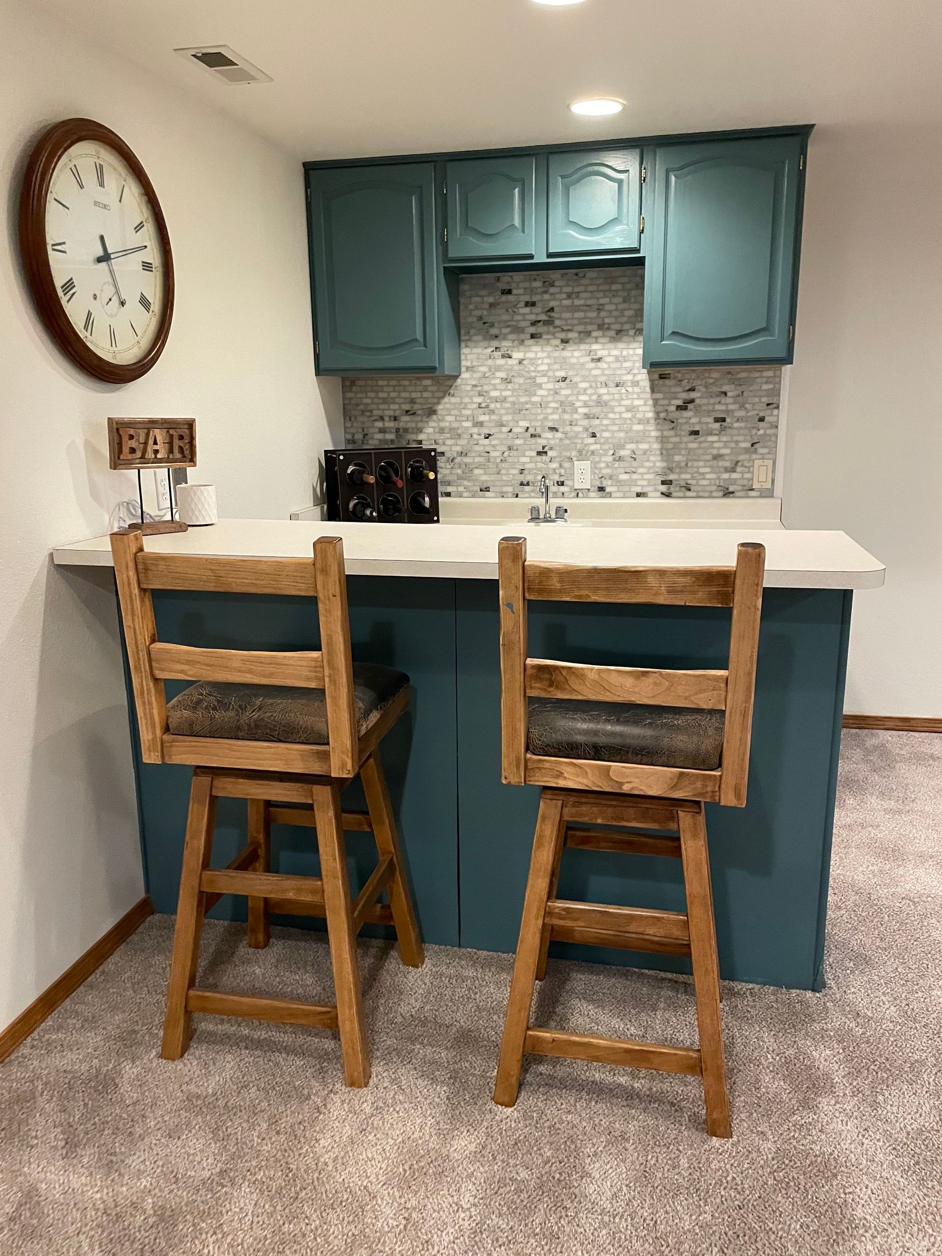 Basement bar with teal cabinets, a white countertop, and two wooden bar stools.