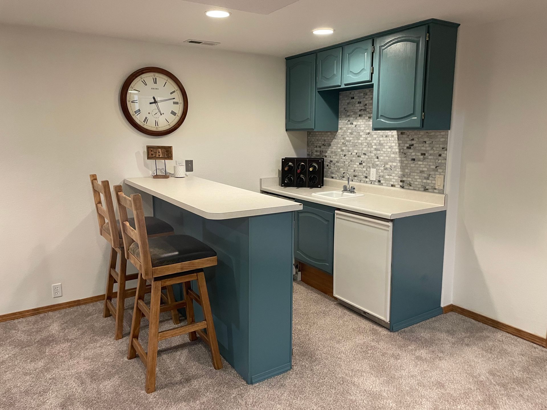 Basement kitchenette with teal cabinets, white countertop, a clock, and stools.
