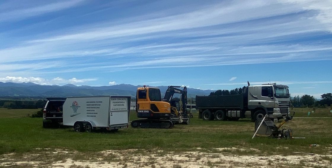 A yellow excavator, a white trailer, and a grey dump truck parked in a grassy field under a bright blue sky.