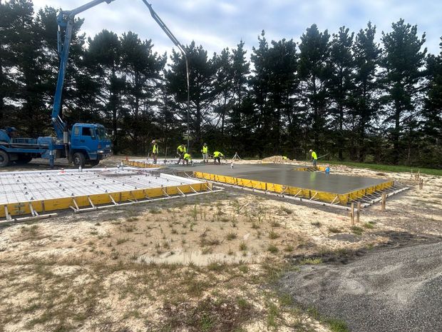 A blue concrete pump truck pours cement into a yellow-framed foundation at a construction site with trees in the background.