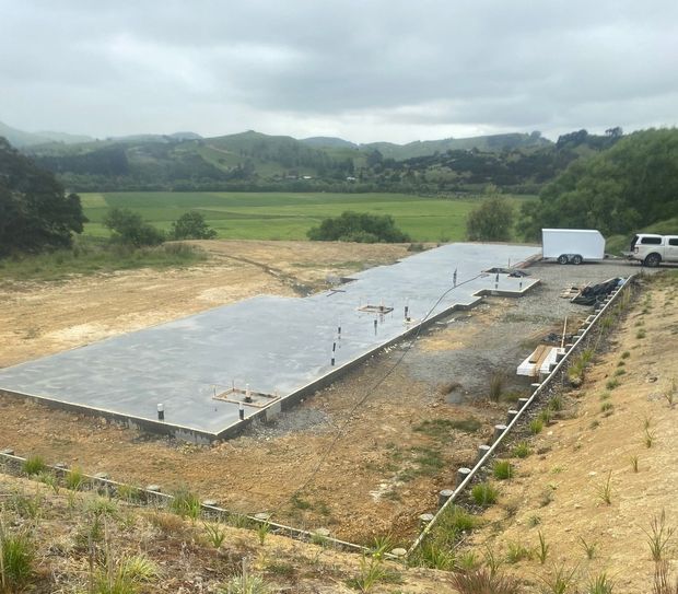 A rectangular concrete foundation for a new building sits on a hillside overlooking a green, rural valley.