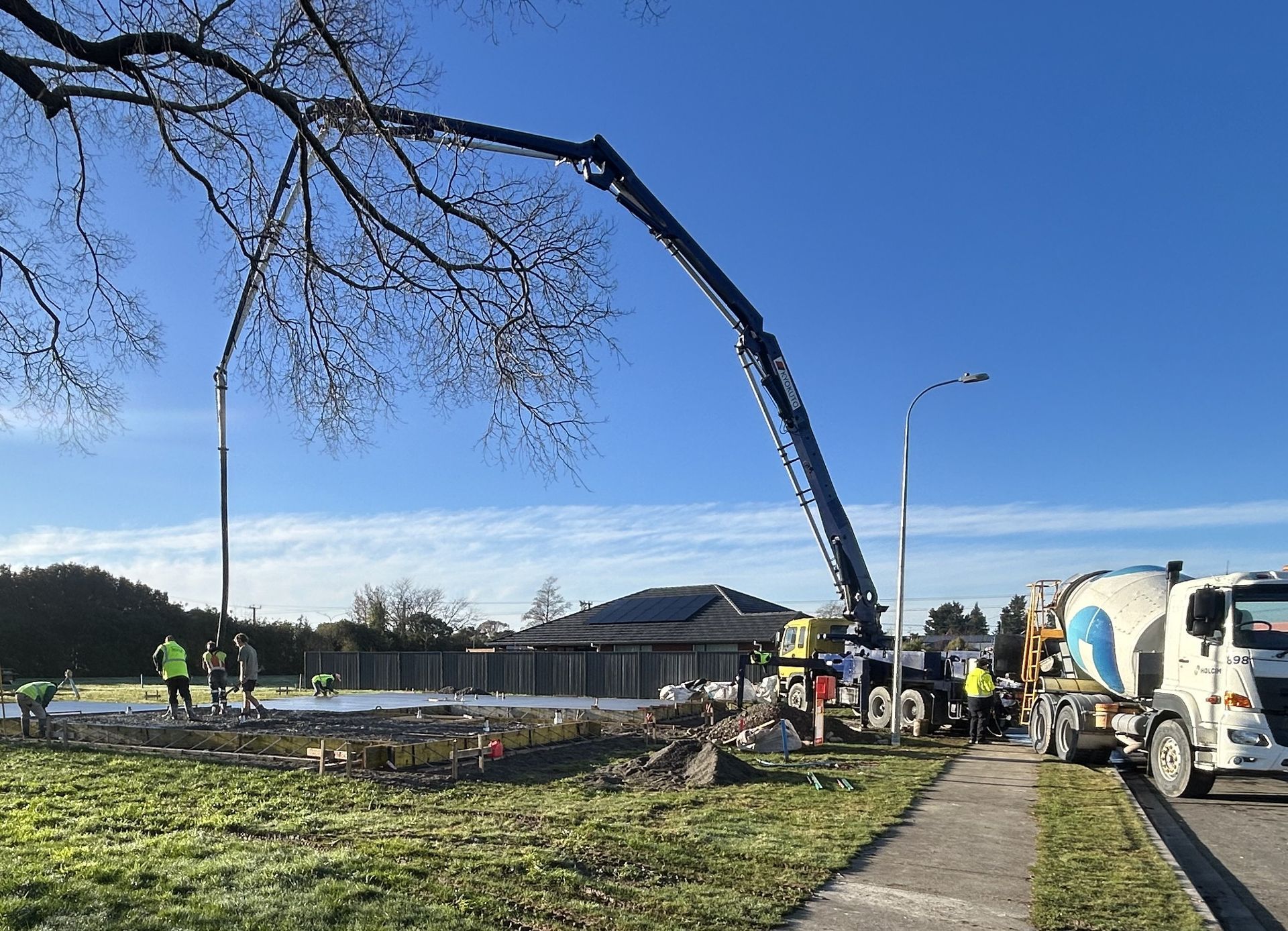 Construction workers use a long concrete pump truck to pour wet cement at an outdoor job site on a sunny day.