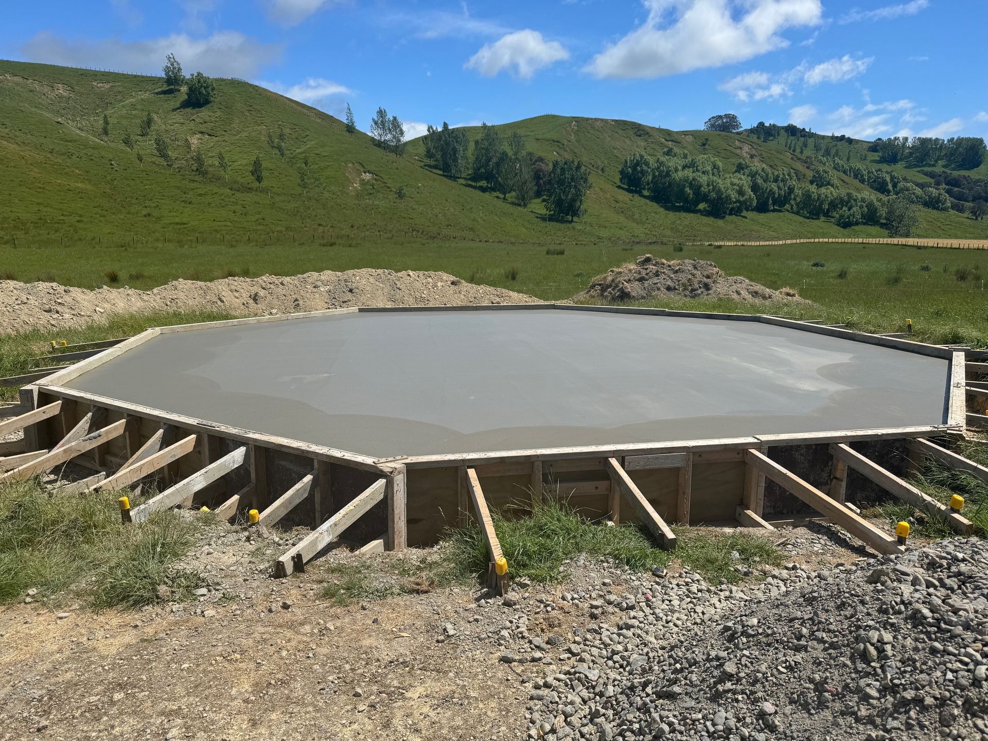 A fresh, octagonal concrete foundation slab poured within wooden formwork on a grassy, hilly construction site.