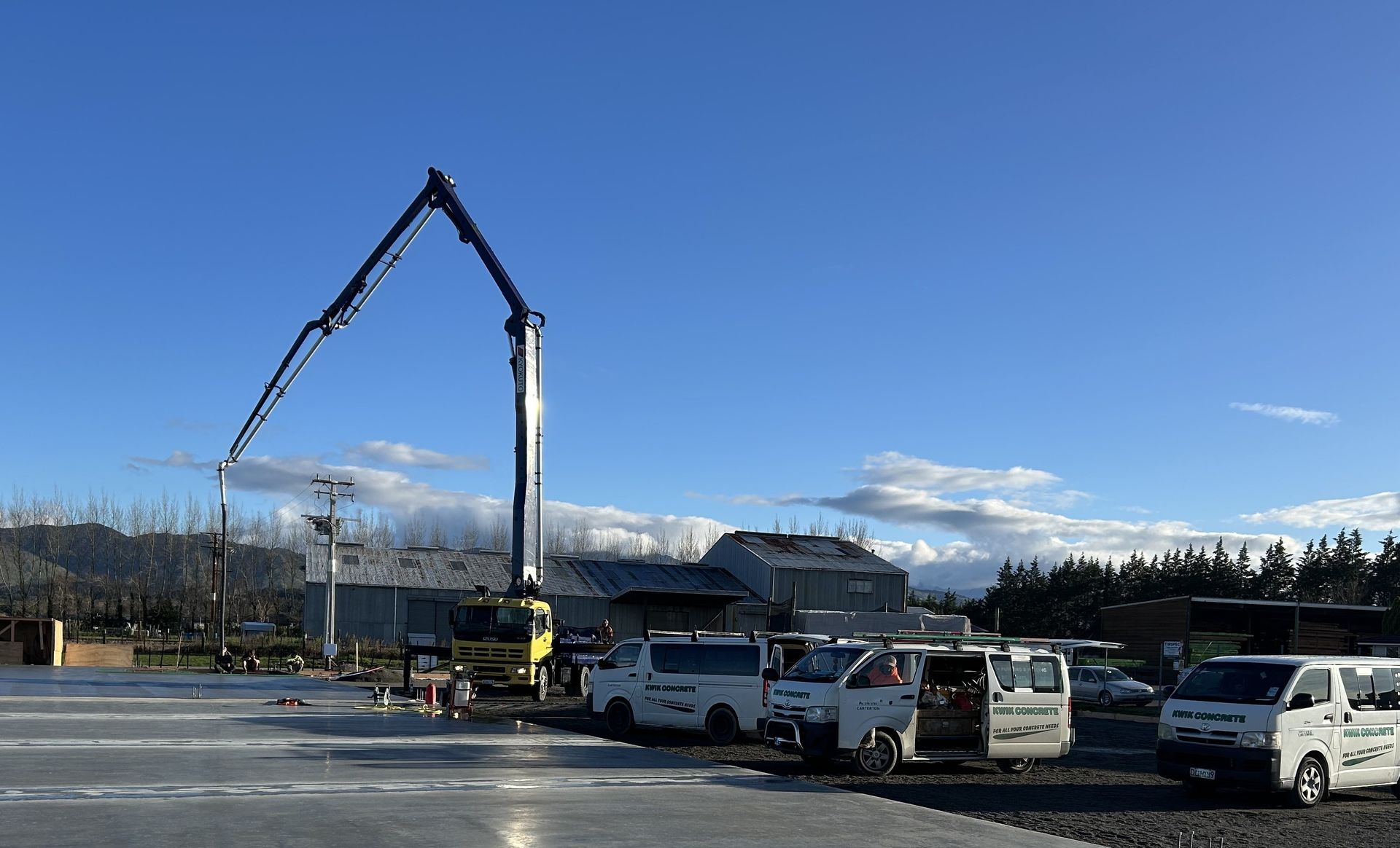 A yellow concrete pump truck extending its arm over a construction site with several white utility vans parked nearby.