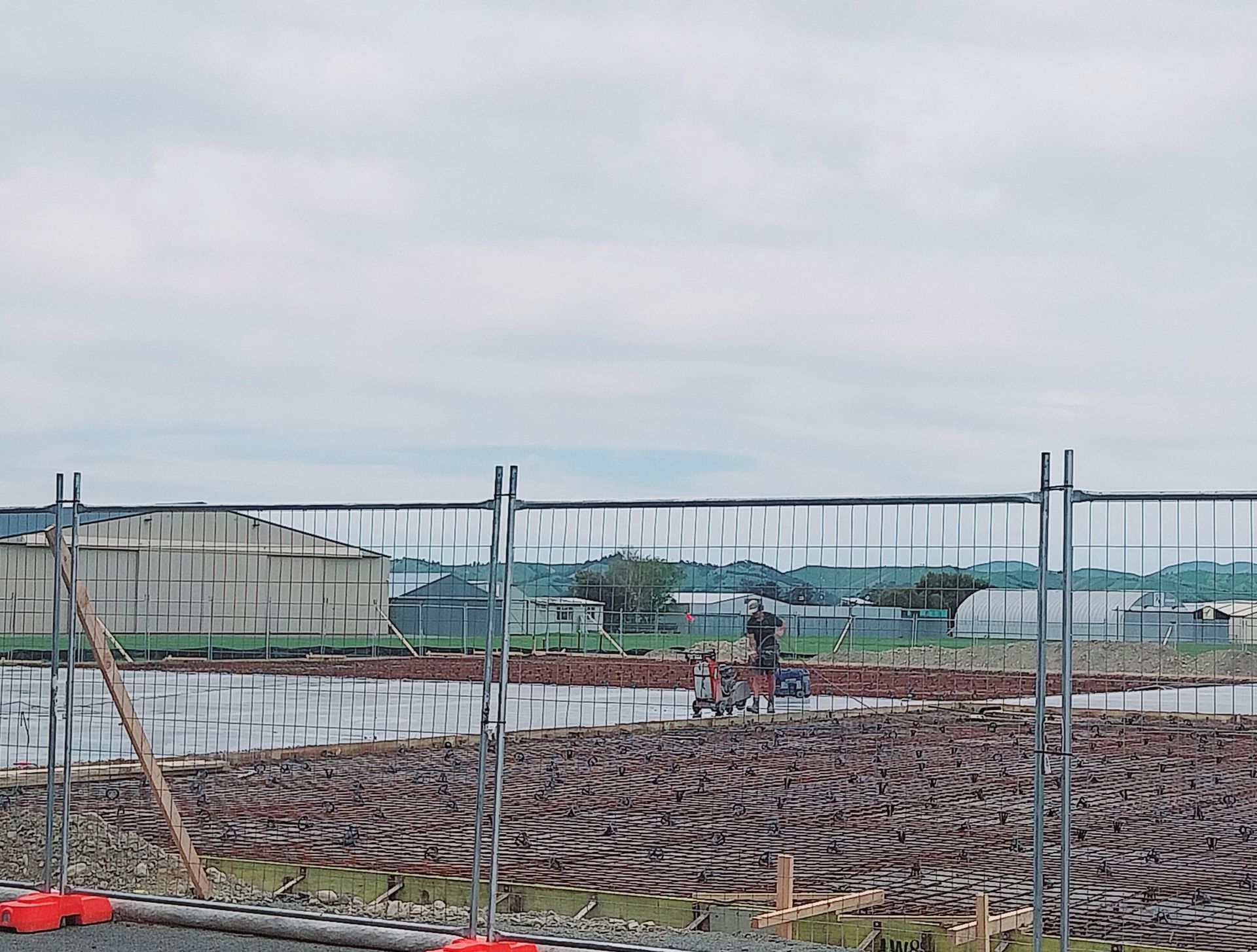 A person stands in a muddy, tilled field behind a chain-link fence, with industrial buildings visible in the distance.