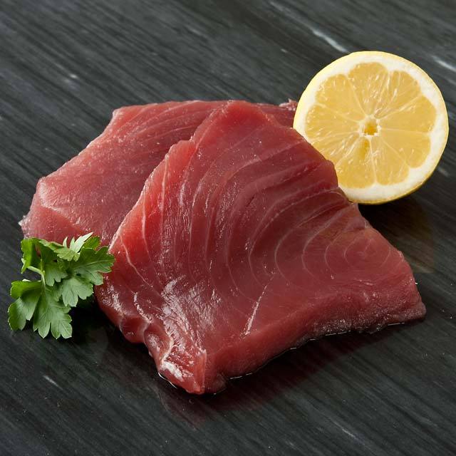 Two raw tuna steaks with a lemon half and parsley on a dark surface.