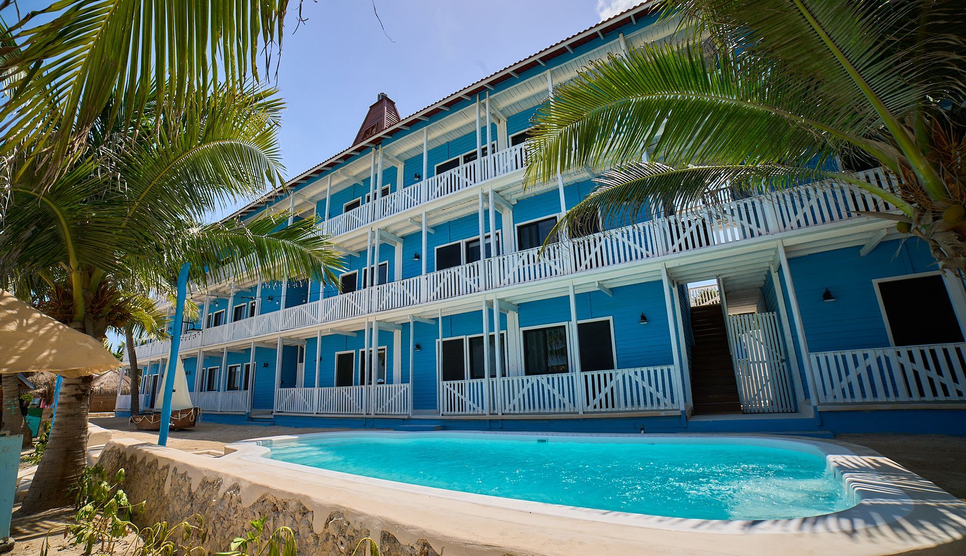 Complejo turístico azul frente a la playa con balcones, palmeras y una piscina bajo un cielo despejado.
