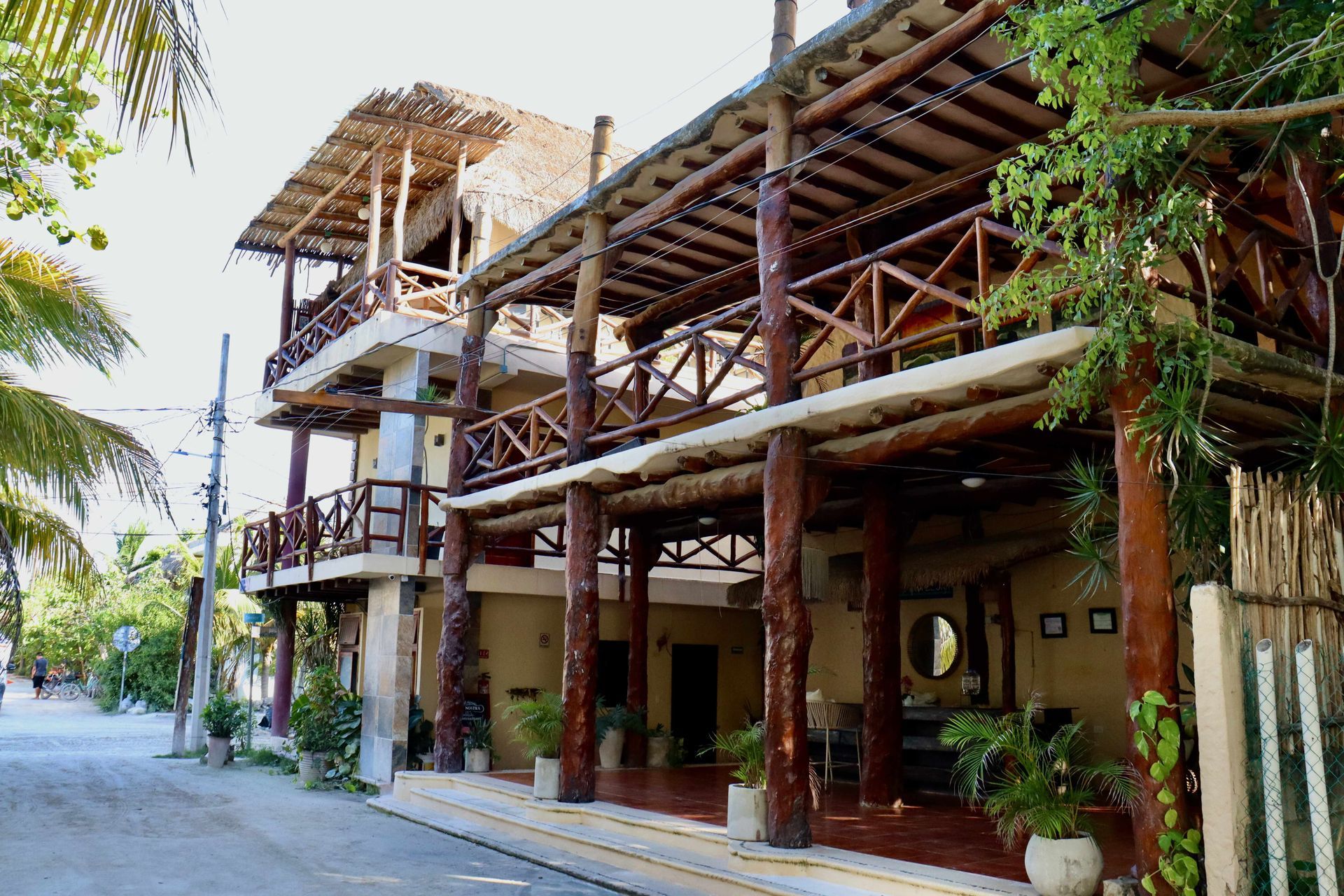 Cabaña de madera con balcones cubiertos y vegetación tropical a lo largo de una calle tranquila.