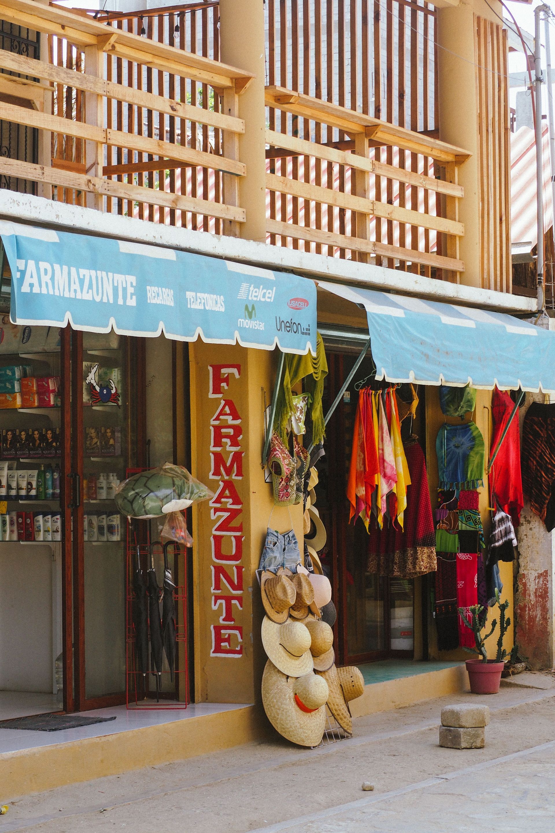 Tienda a pie de calle con toldo azul, ropa colgada y un cartel con un osito de peluche en la entrada.