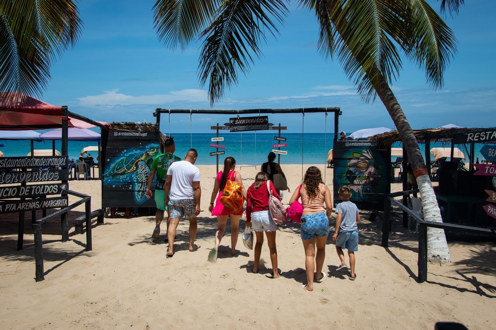 Gente caminando hacia la entrada de una playa con palmeras, arena y un océano turquesa al fondo.
