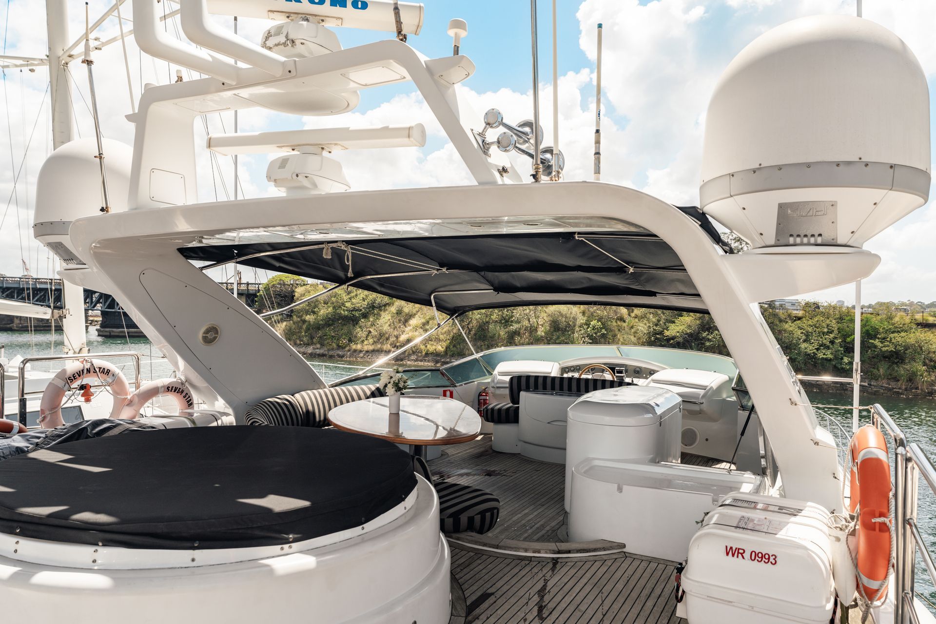 Yacht deck with a long, striped sofa, overlooking other boats in a marina, under a cloudy blue sky.