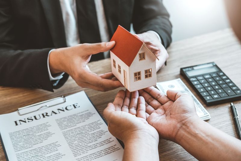 Hands holding a small house model over insurance paperwork with a calculator nearby