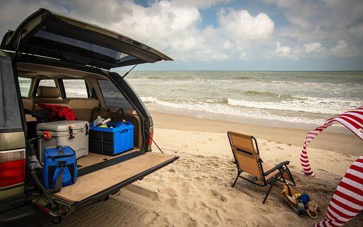 Open SUV with beach gear parked on a sandy shore beside ocean waves and a striped umbrella