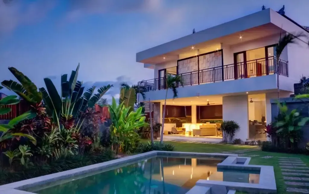Modern two-story villa with lit patio, tropical landscaping, and a swimming pool at dusk