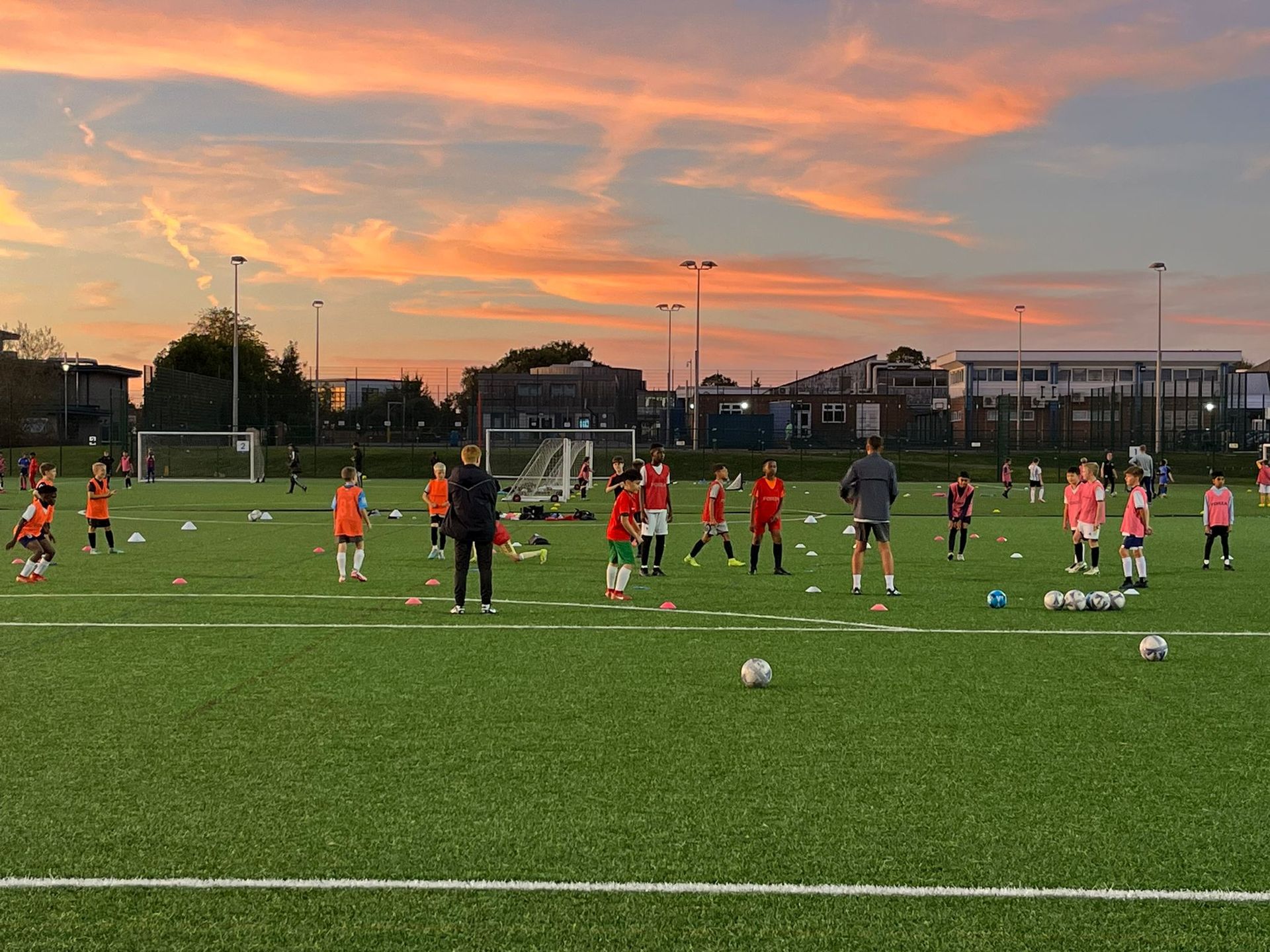 Children practicing football on a field at sunset, with coaches and balls scattered on the grass.