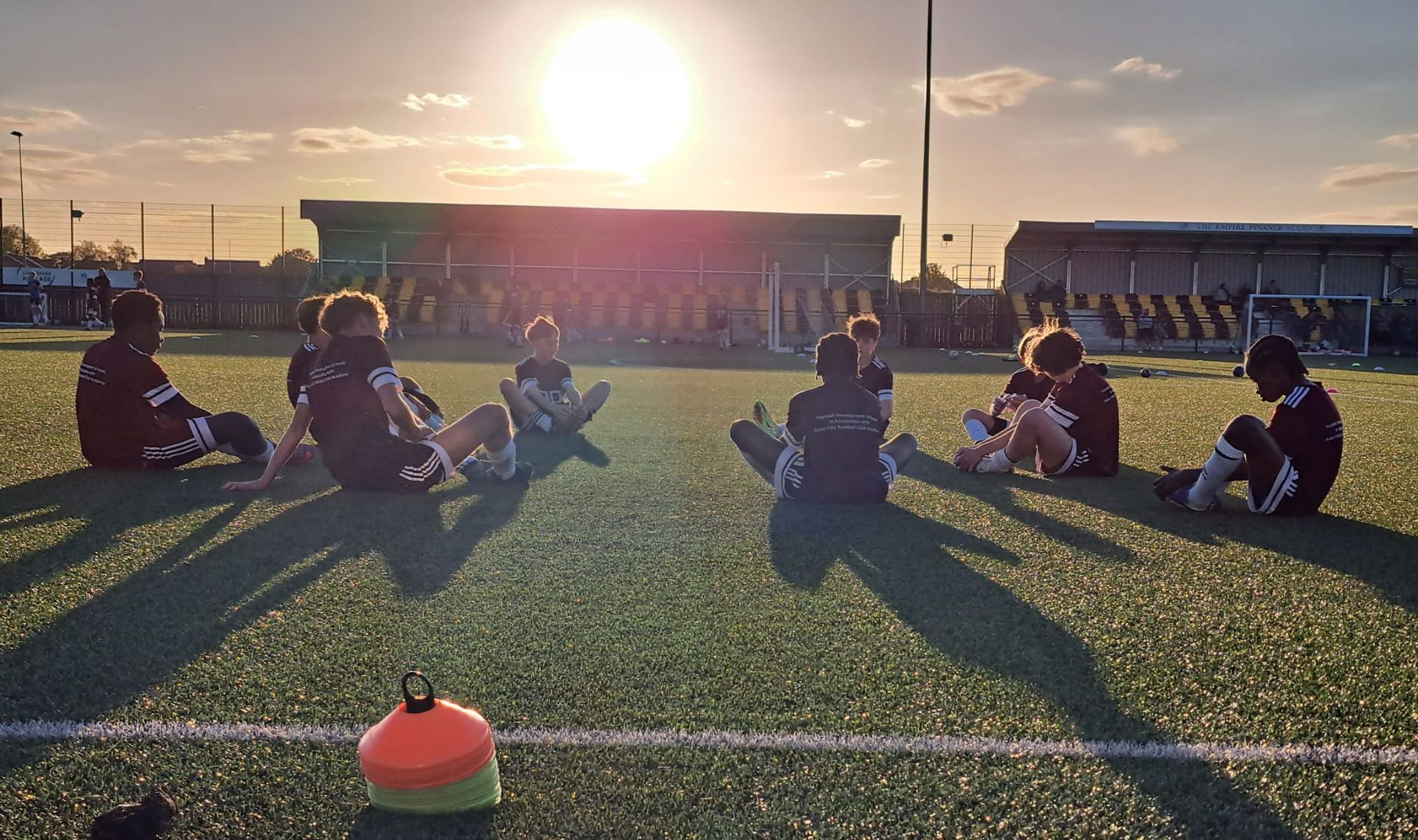 Players sitting on a sports field at sunset, with long shadows and a bright sun low on the horizon.