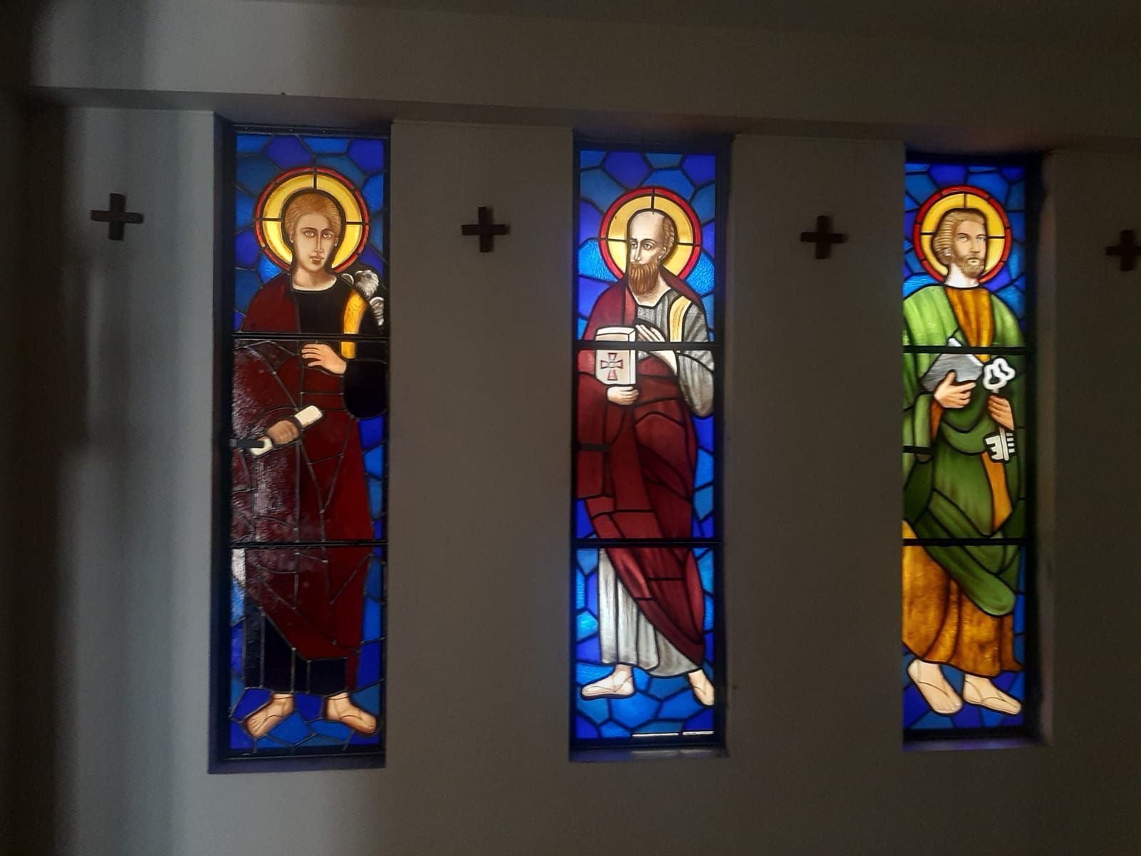 Vitral que muestra tres figuras vestidas con túnicas azules, rojas y doradas dentro de una iglesia de piedra.