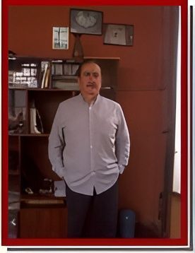 Hombre de pie en el interior de una habitación con paredes rojas, vistiendo una camisa clara abotonada y con las manos en los bolsillos.