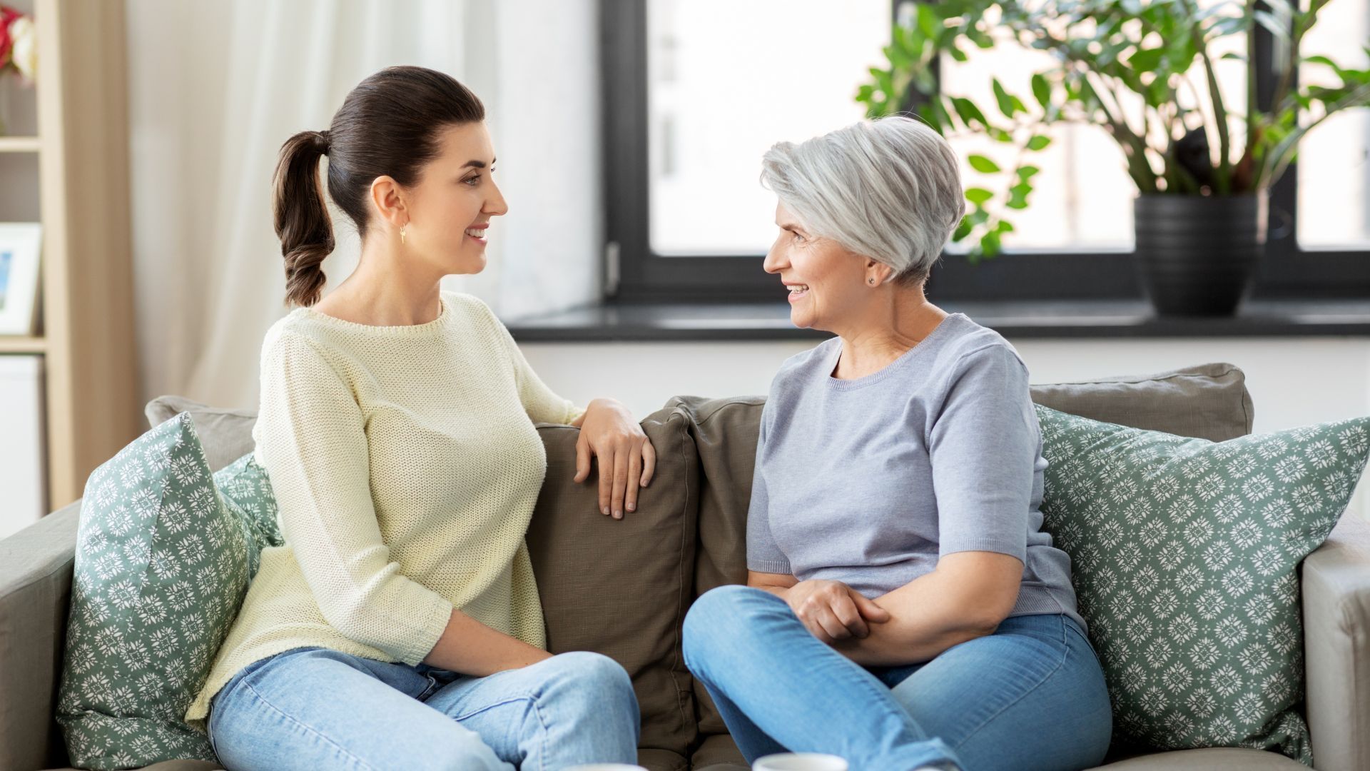 Woman in ponytail talks to an older woman with gray hair on a couch indoors; smiles, light.