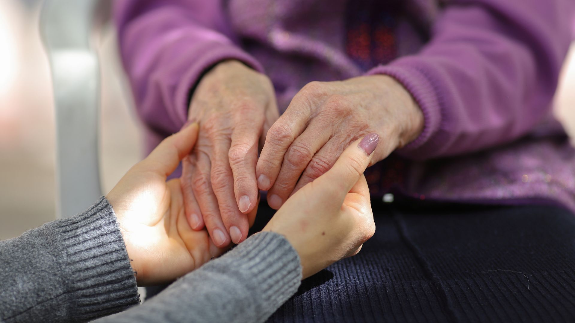 Person holding the hands of an older person. Purple sweater, light skin, supportive gesture.
