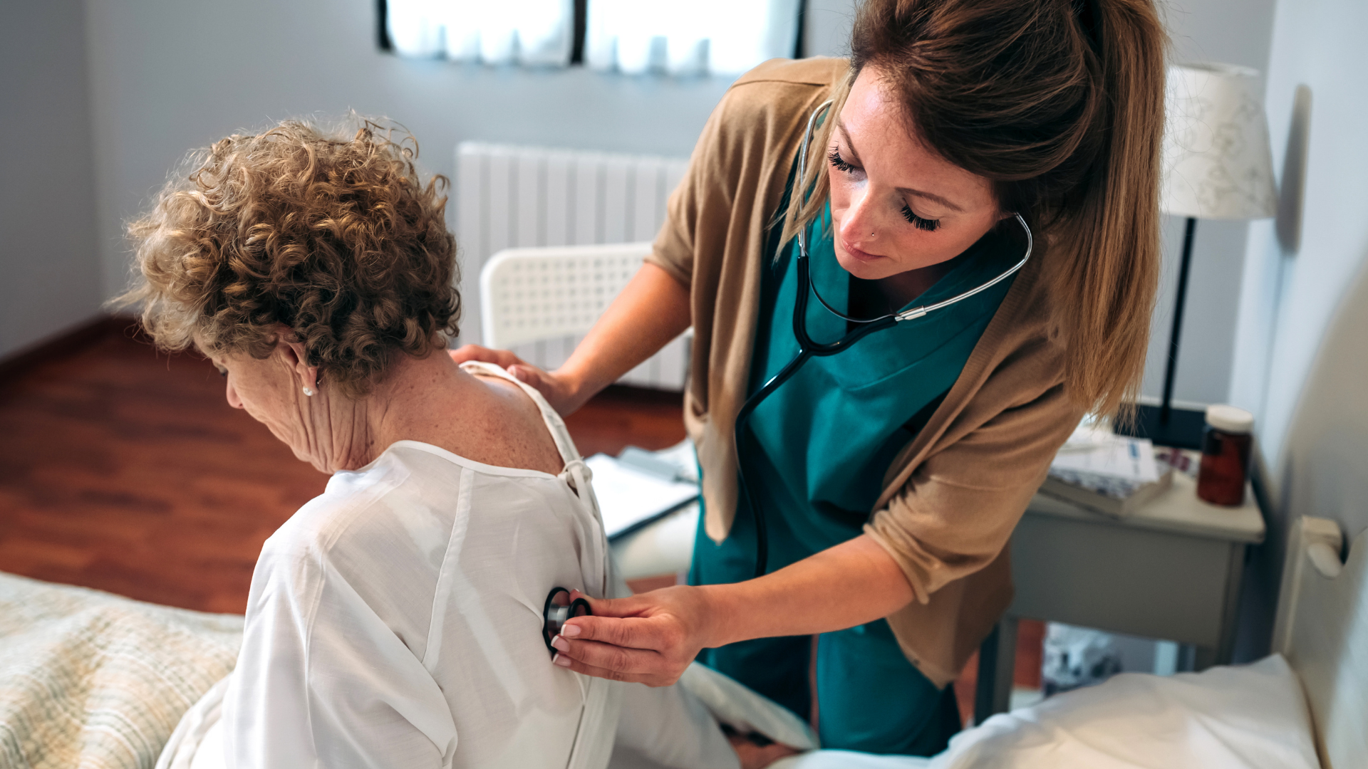 A nurse listens to an elderly woman's back with a stethoscope in a bedroom setting.