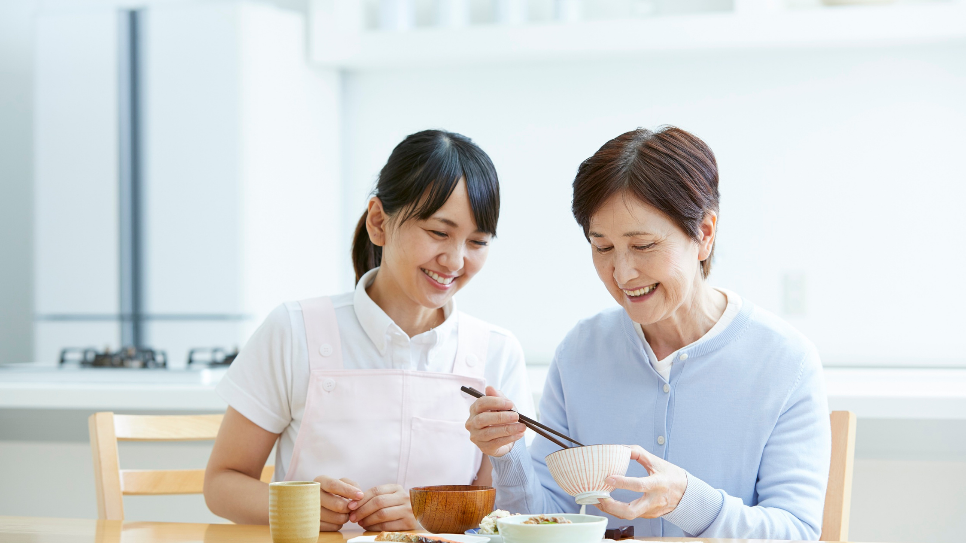 Woman helping senior eat with chopsticks at a table; smiles, light setting.