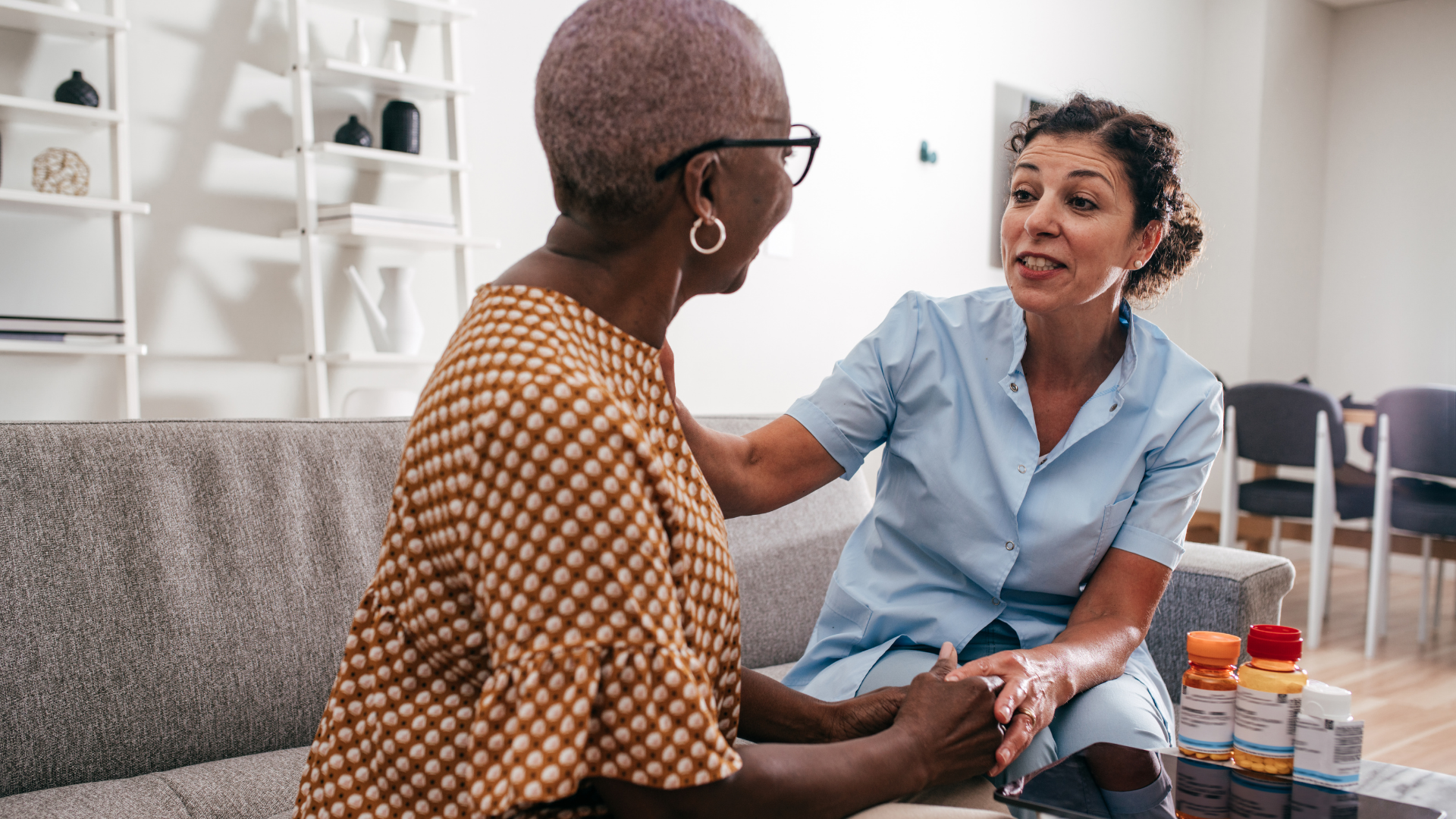 Caregiver with light skin, in blue scrubs, comforts elderly Black woman on a couch, who wears glasses and patterned top.