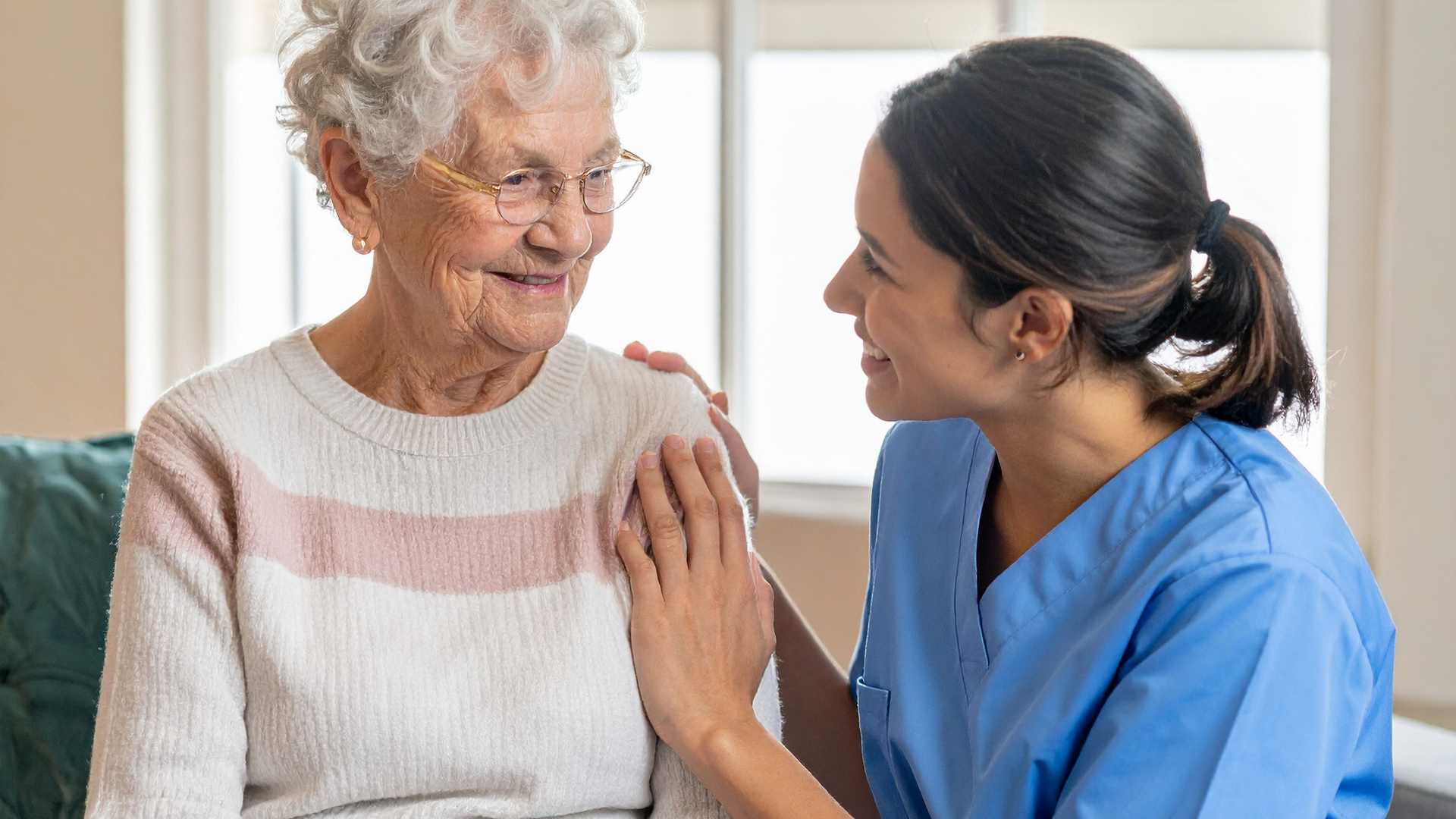 Older woman smiles as a caregiver places a hand on her shoulder indoors.