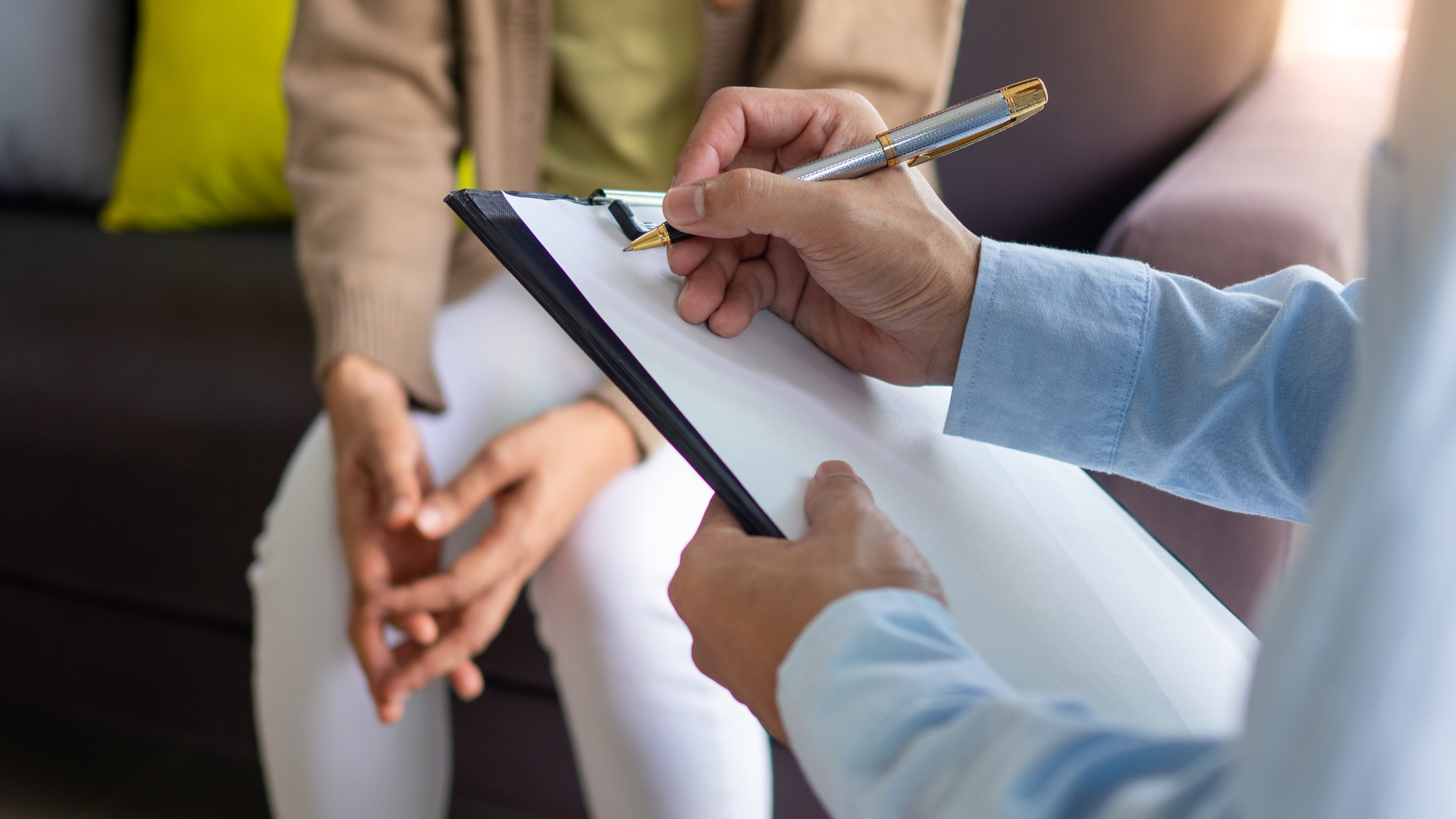 A person sits on a couch as another person takes notes on a clipboard.
