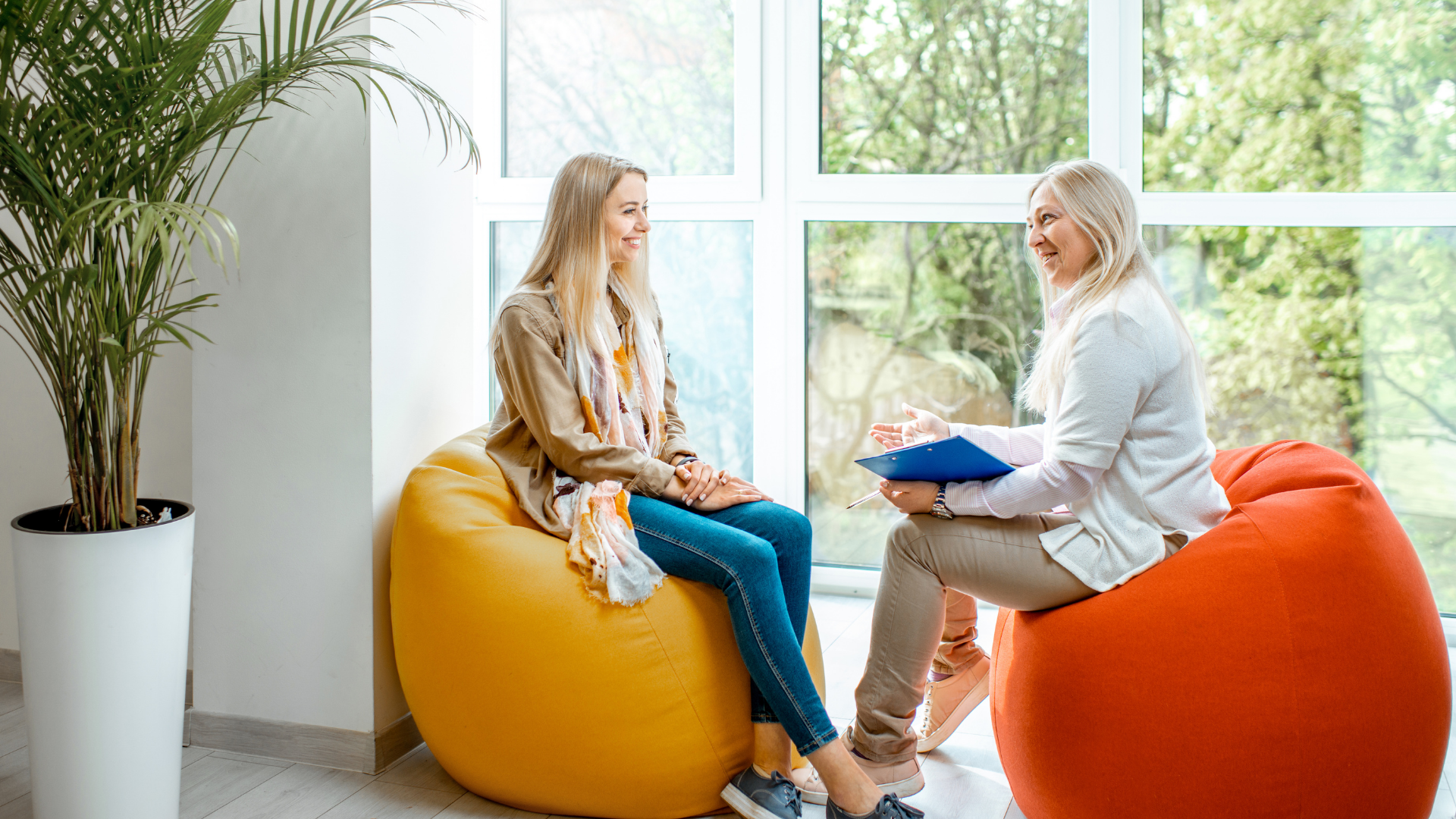 Woman in therapy session, sitting on beanbags, smiling, talking to a therapist by a window.