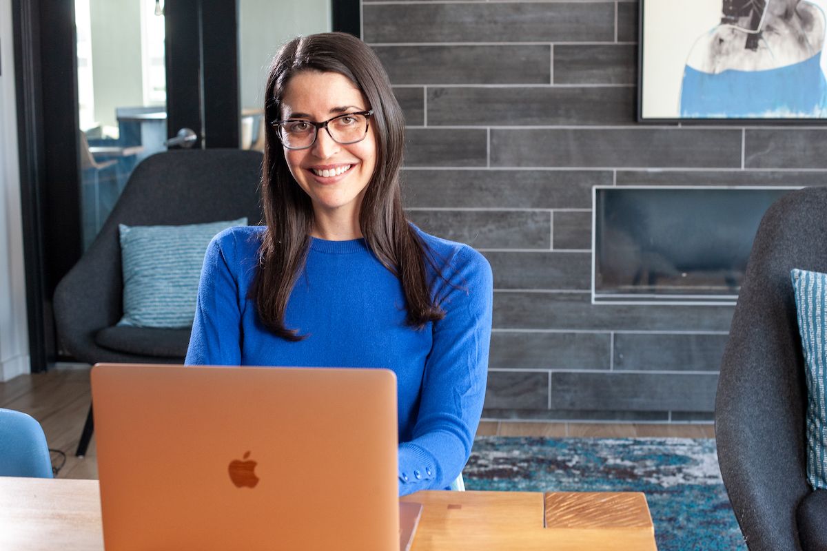 Woman with dark hair and glasses smiles, wearing a yellow sweater. She sits in an office setting.