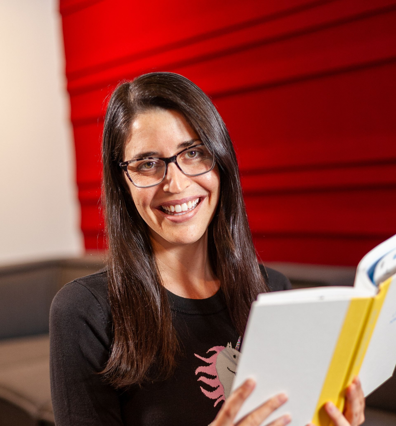 Woman with glasses smiles, reading a book on a couch, red wall behind her.