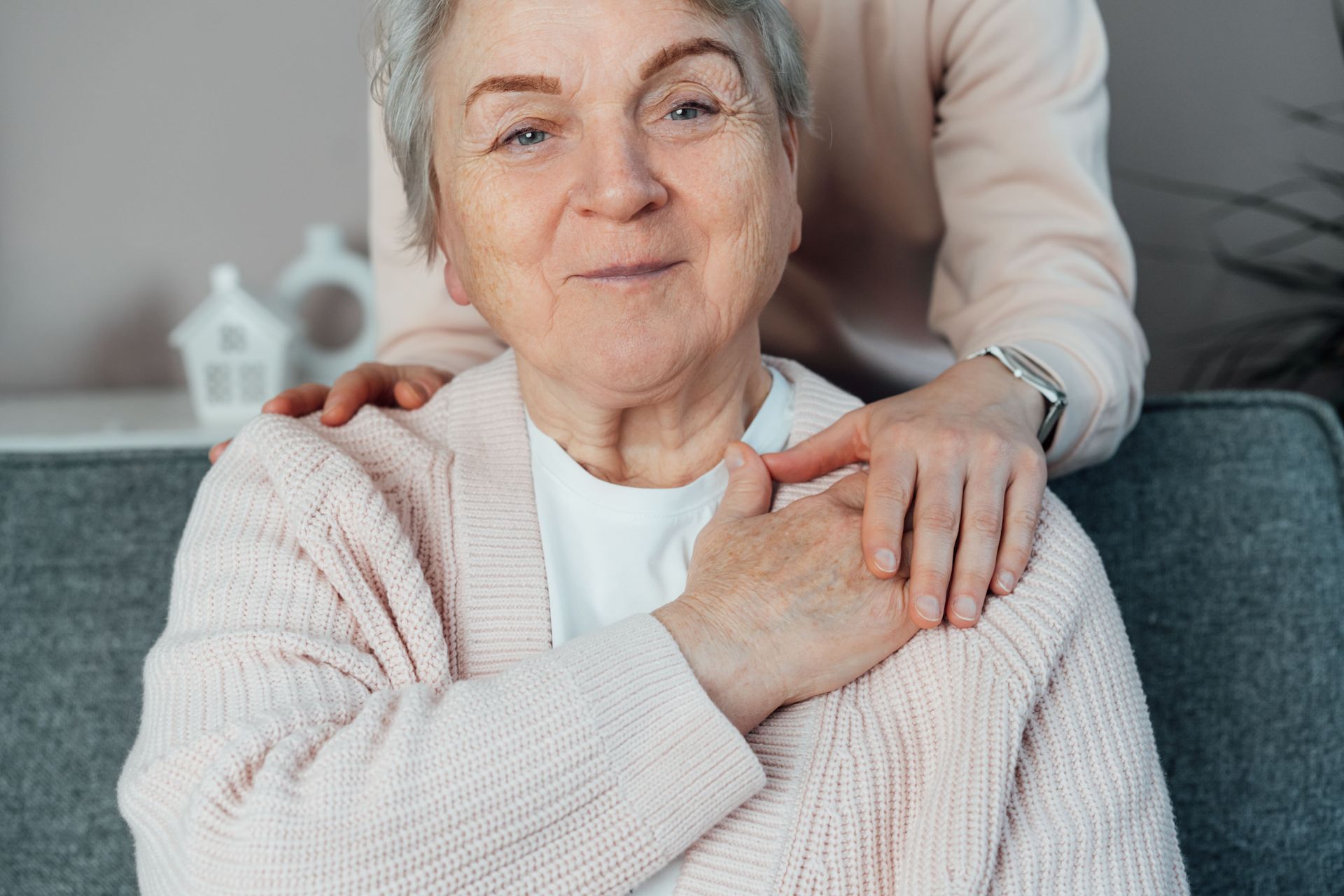 Woman with ponytail assisting an elderly man drawing at a table, smiling. Sunlight streams through window.