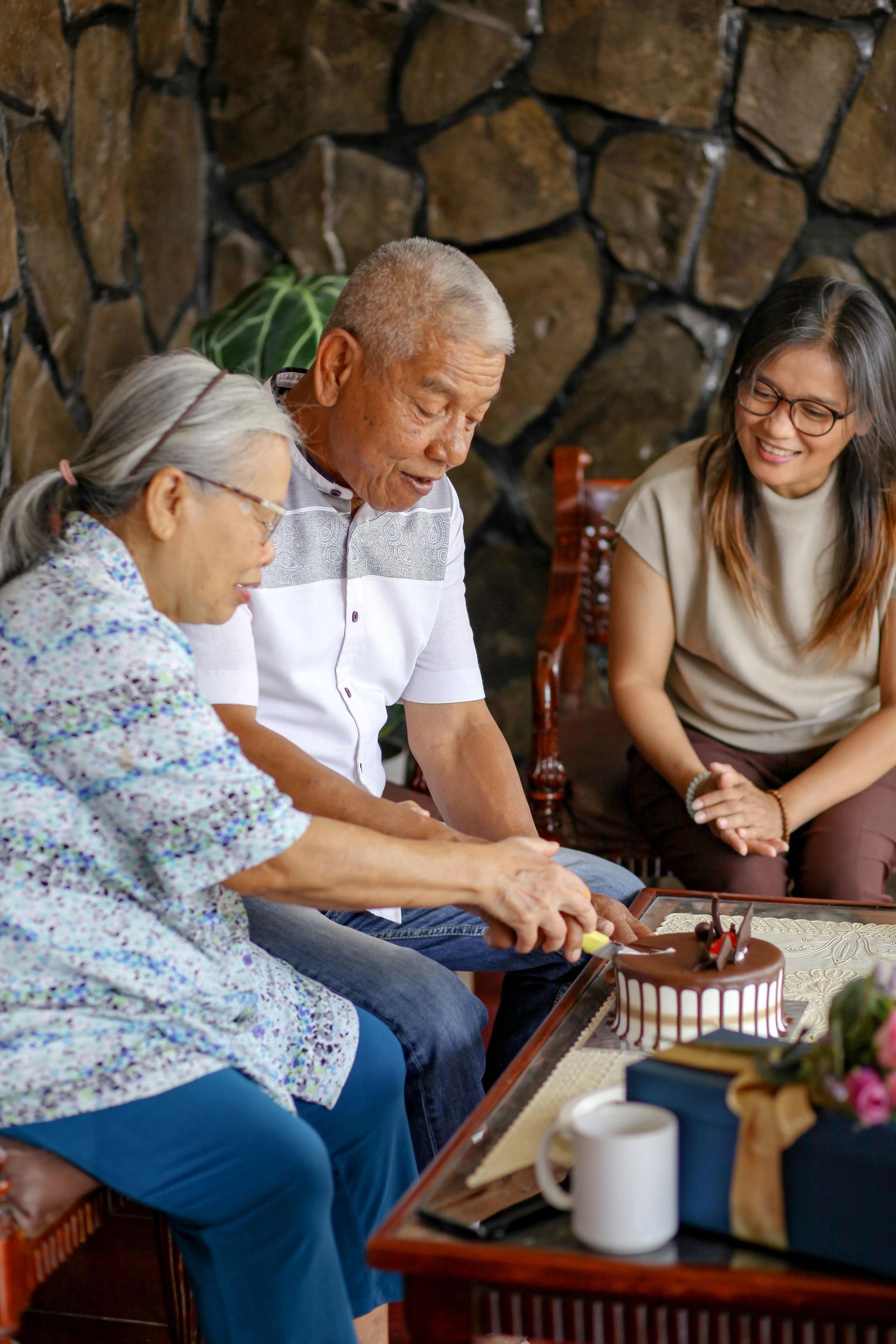 An older man sits at a table, looking down; a woman prepares something beside him.